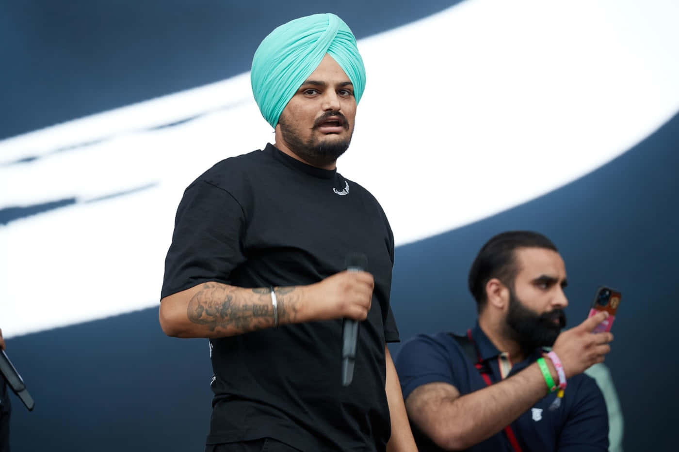戴著藍綠色頭巾的男子在戶外活動中對著麥克風講話。 — Sidhu Moose Wala 壁紙