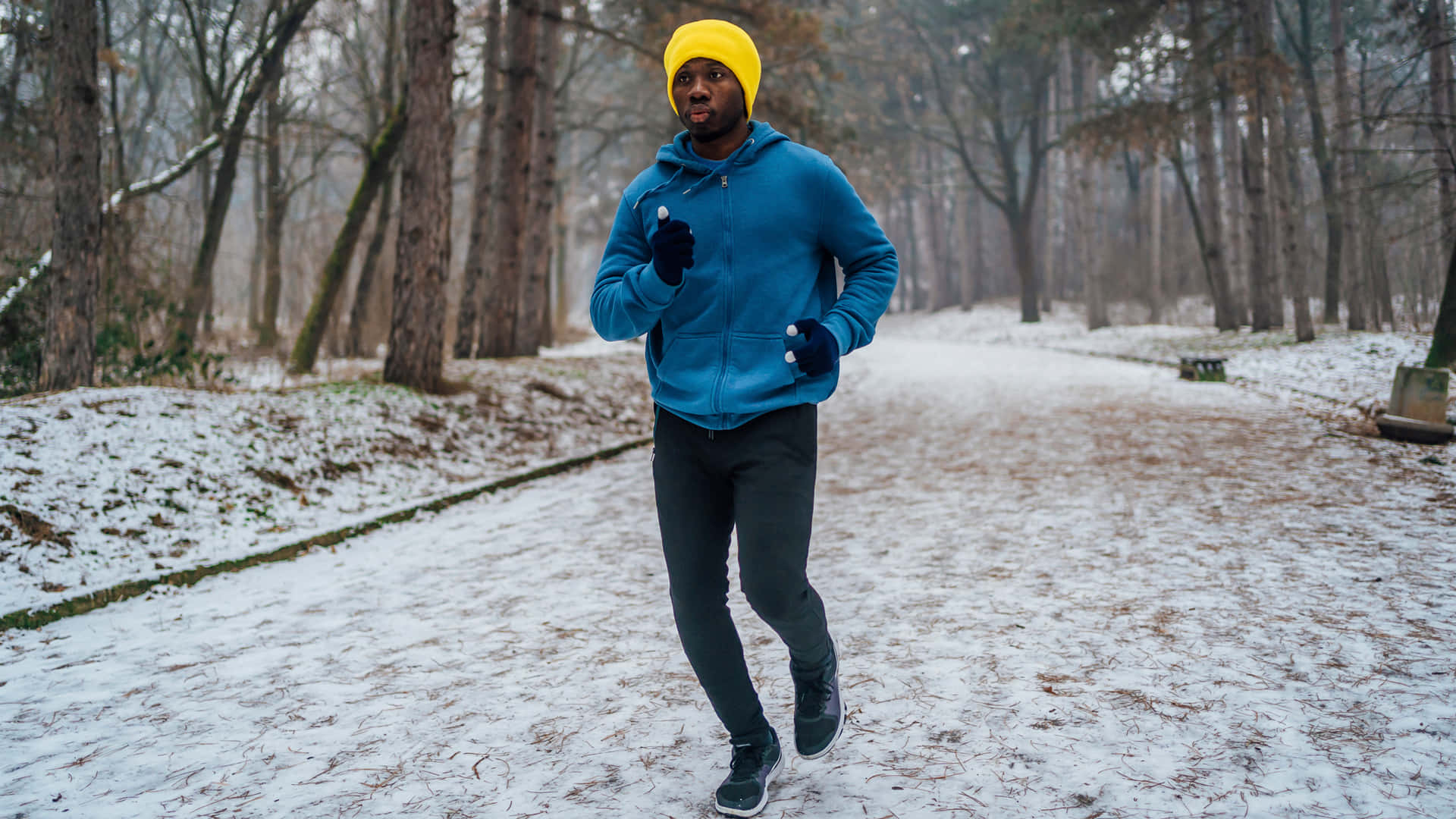 Running 桌布 壁紙 — 一名戴著亮黃色毛帽、身穿藍色外套的男子，在兩旁光禿樹木的積雪森林小徑上奔跑。