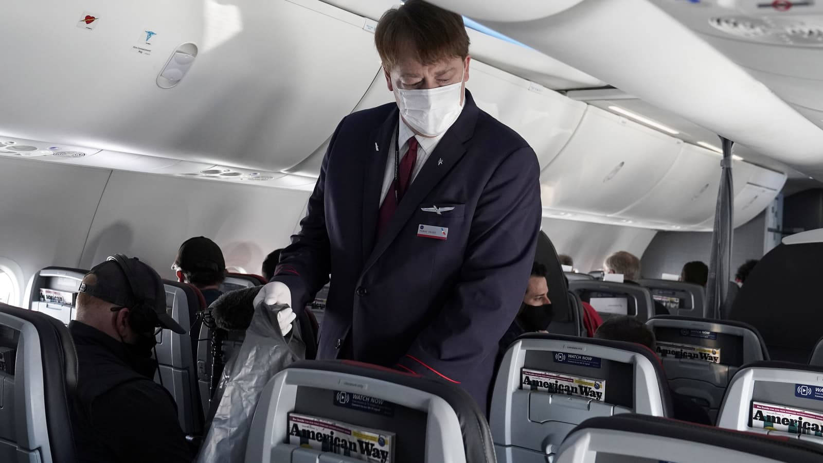 Airport 桌布 壁紙 — 一名戴著口罩、身穿深色制服的空中服務員在美國航空公司的飛機上為乘客提供服務，可見 overhead bins 和座椅背螢幕。