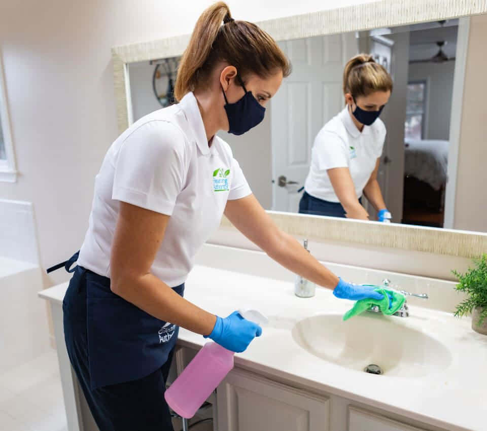 Cleaning 桌布 壁紙 — 一名戴著口罩、身穿白色制服的清潔工，戴著藍色手套，用粉紅色瓶子噴灑和擦拭浴室洗手盆。