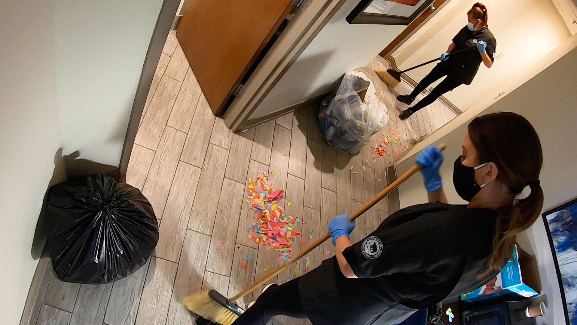 Work 桌布 壁紙 — 一名戴著口罩和手套的清潔工正在掃走木地板上的彩色紙屑，而另一名工人則在走廊鏡子中映照她的動作。