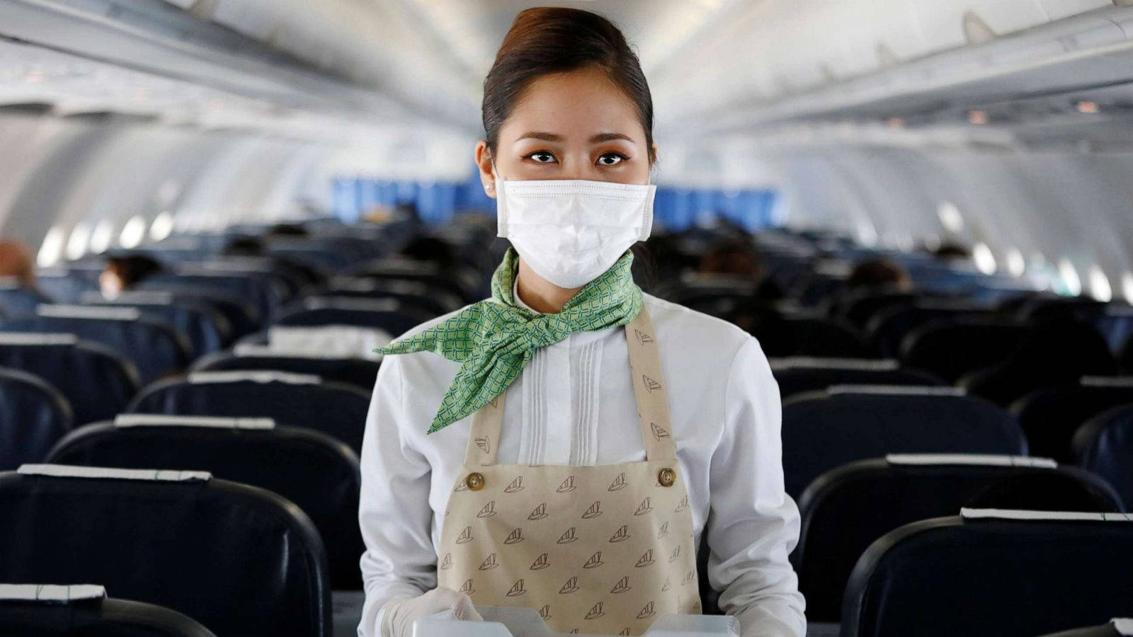 飛機 桌布 壁紙 — 一名戴著口罩和綠色圍巾的空中服務員站在空蕩蕩的飛機艙內，艙內有一排排深藍色的座位。