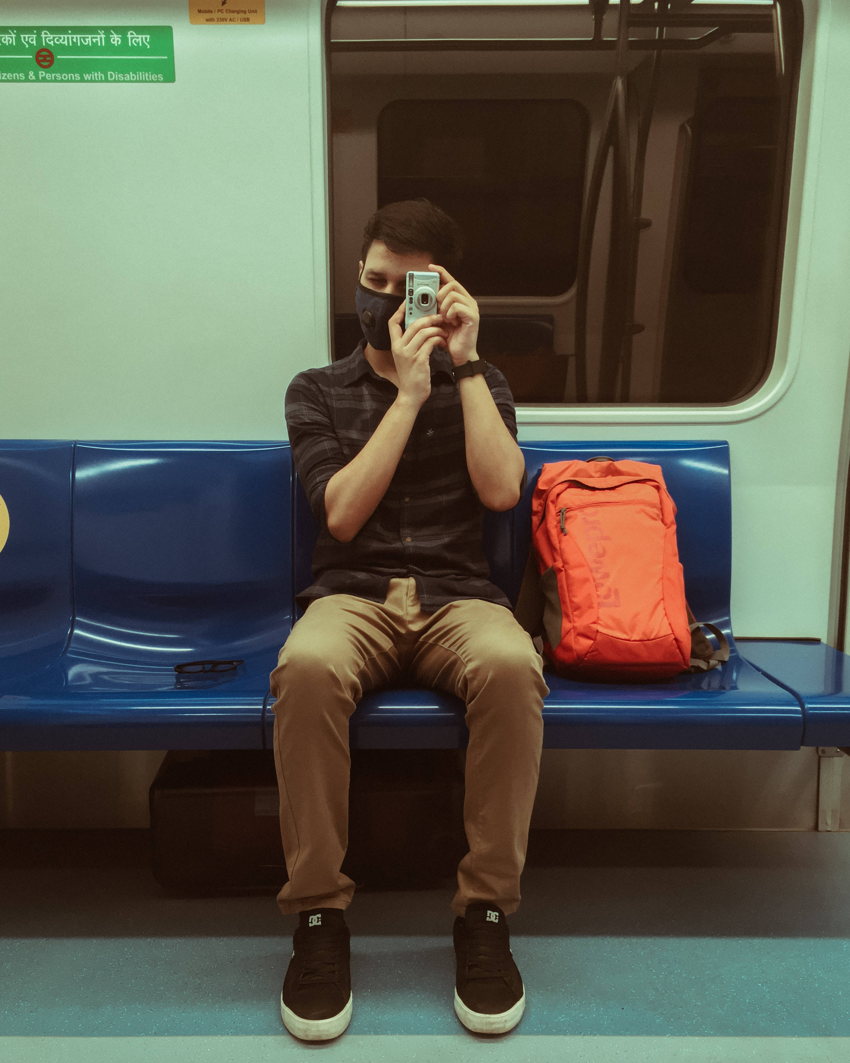 Man Aesthetic 桌布 壁紙 — 一名戴著口罩的人坐在藍色地鐵長椅上，拿著相機自拍，旁邊有一個橙色背包。