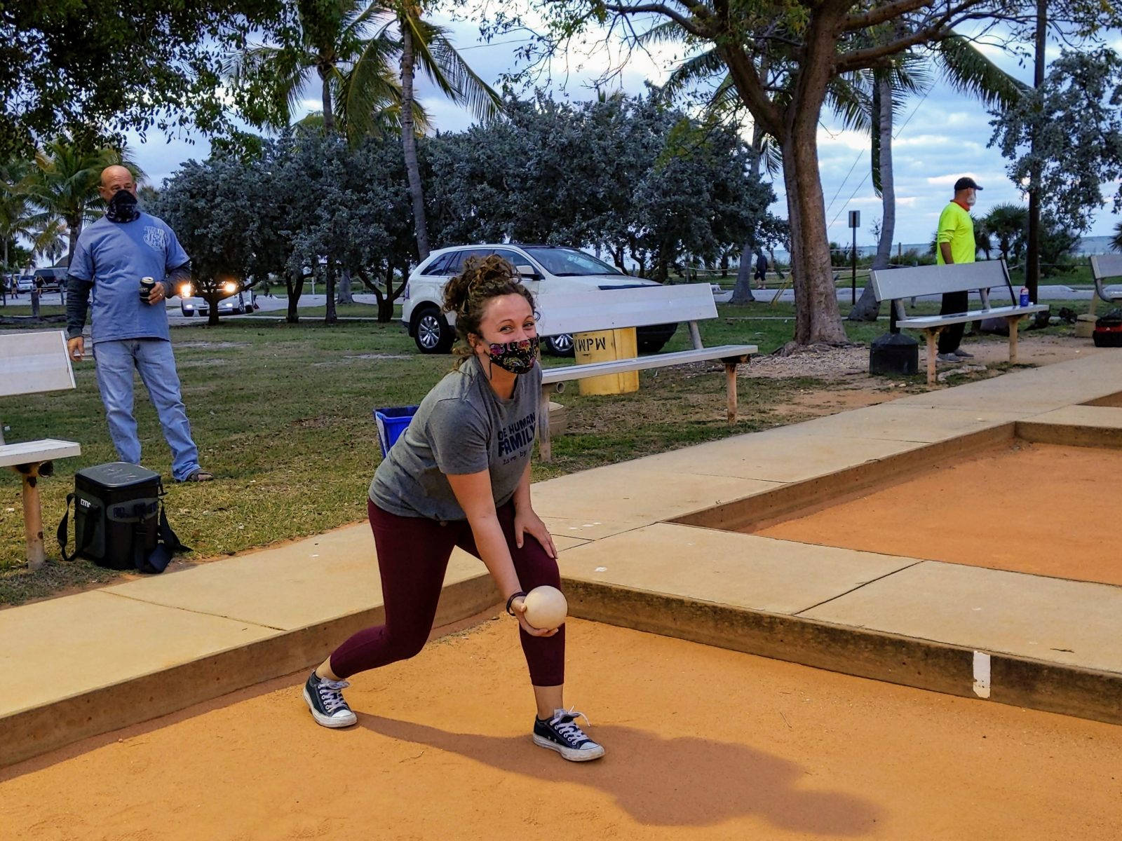 運動 桌布 壁紙 — 一名戴著口罩的女子準備在公園擲球，其他人觀看，汽車駛過。