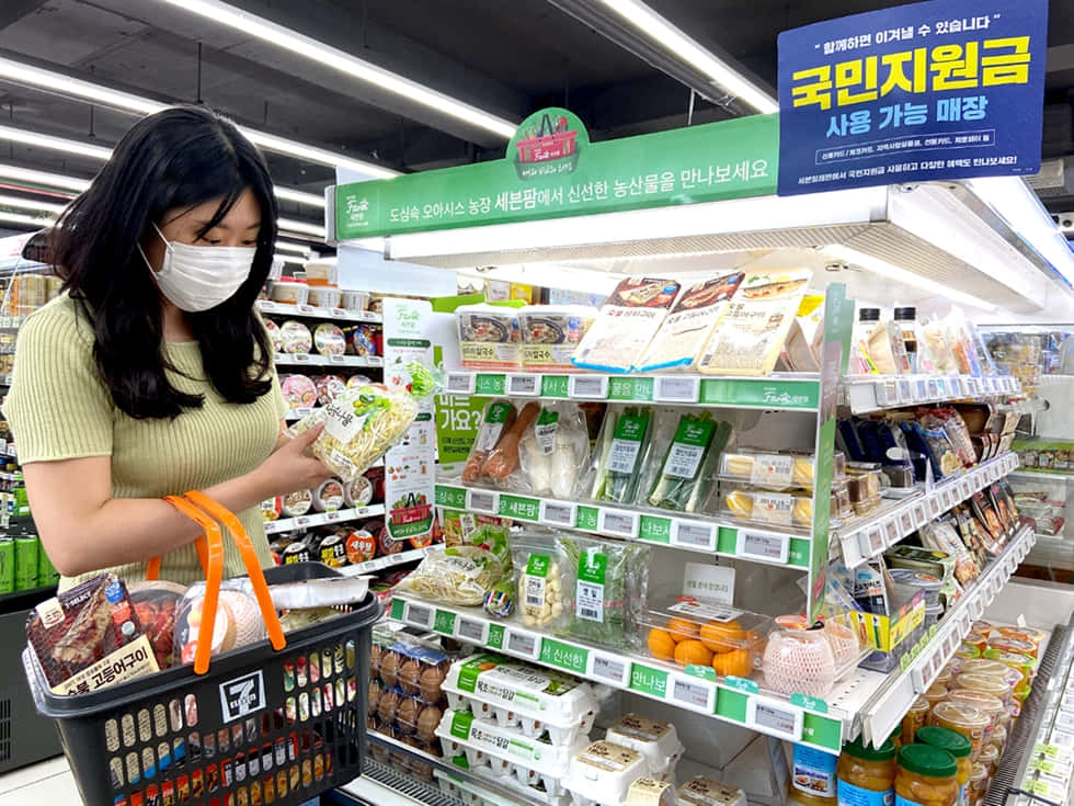 7 Eleven 桌布 壁紙 — 一名戴著口罩的女性在光線明亮的韓國超市通道中瀏覽包裝蔬菜。