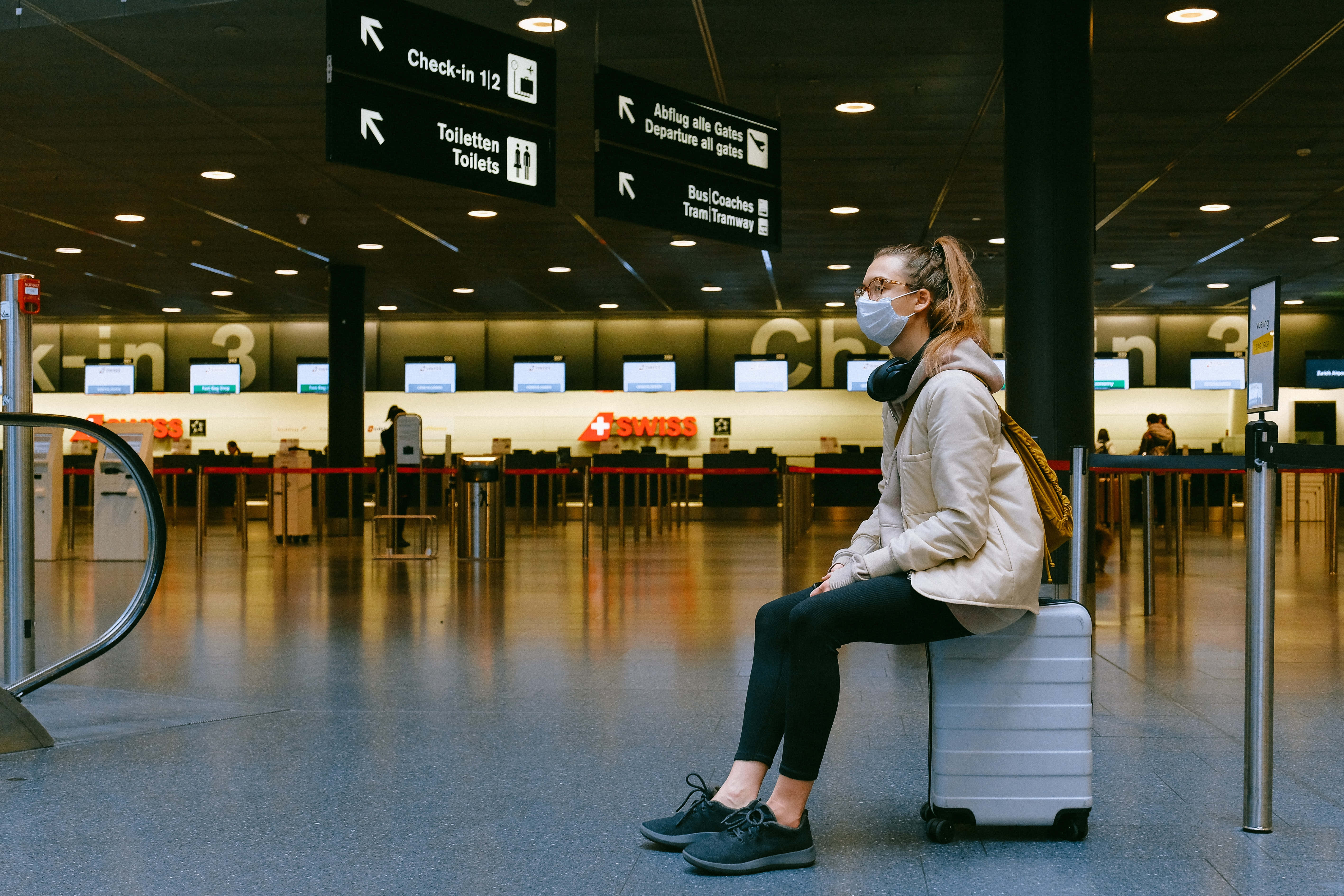 Waiting 桌布 壁紙 — 一名戴著口罩的旅客獨自坐在一個寬敞的機場航廈內的行李箱上，頭頂有辦理登機手續櫃檯和方向指示牌。