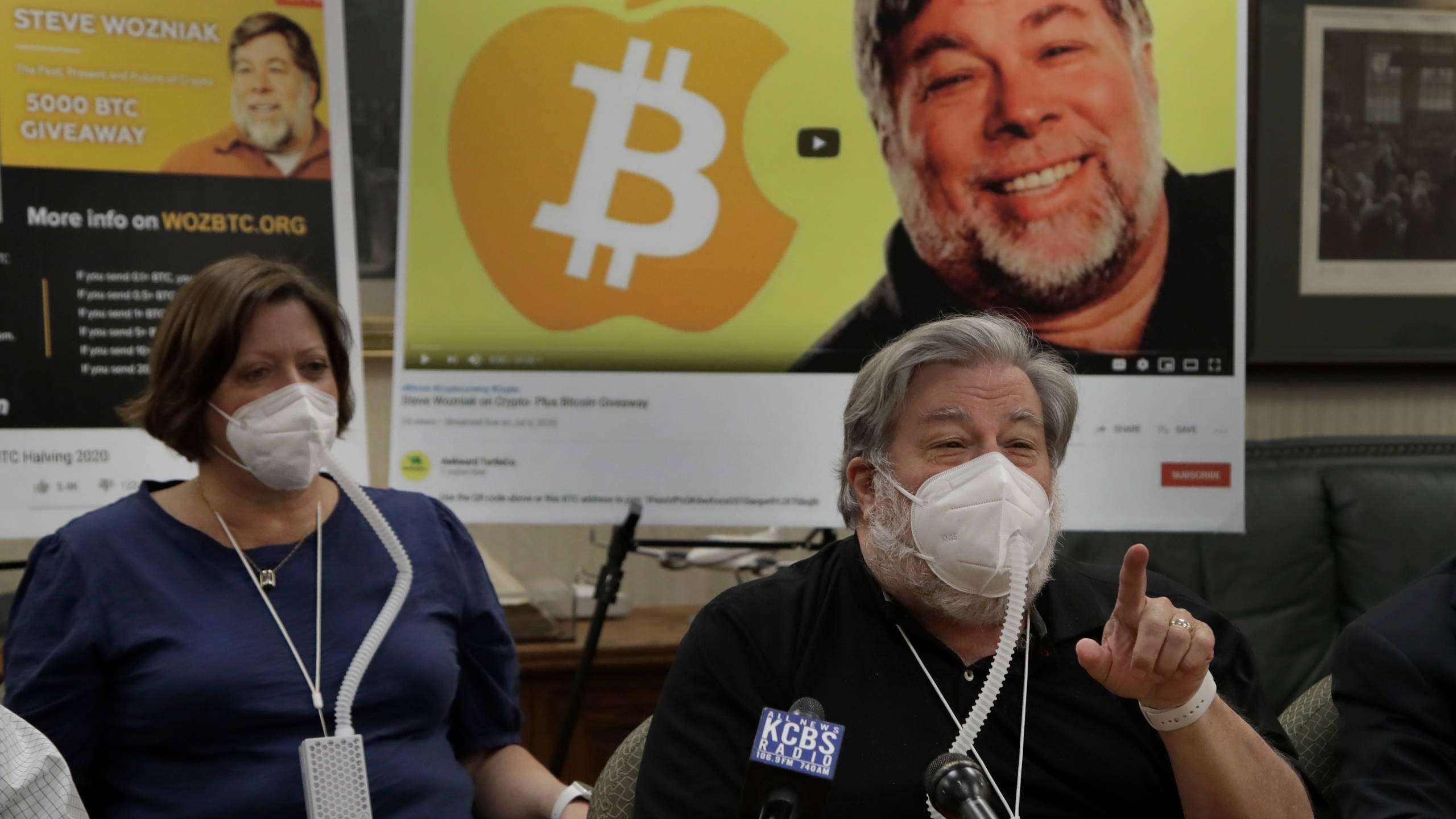 比特幣 桌布 壁紙 — 一名戴著口罩的男子在記者會上坐著做手勢，身後有比特幣宣傳海報。