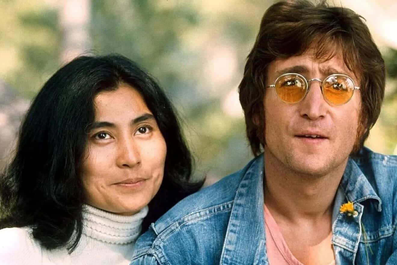 Famous 桌布 壁紙 — 一名戴著圓形橙色鏡片眼鏡及牛仔外套的男子，坐在一位身穿白色高領毛衣的女子旁邊，背景是柔和的綠色模糊效果。