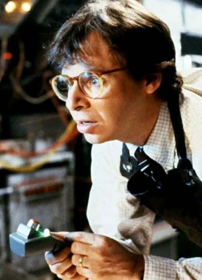 rick moranis 桌布 壁紙 — 一名戴著圓框眼鏡、身穿格紋襯衫的男子，在一個佈滿電線和設備的凌亂實驗室中手持一個發光裝置。