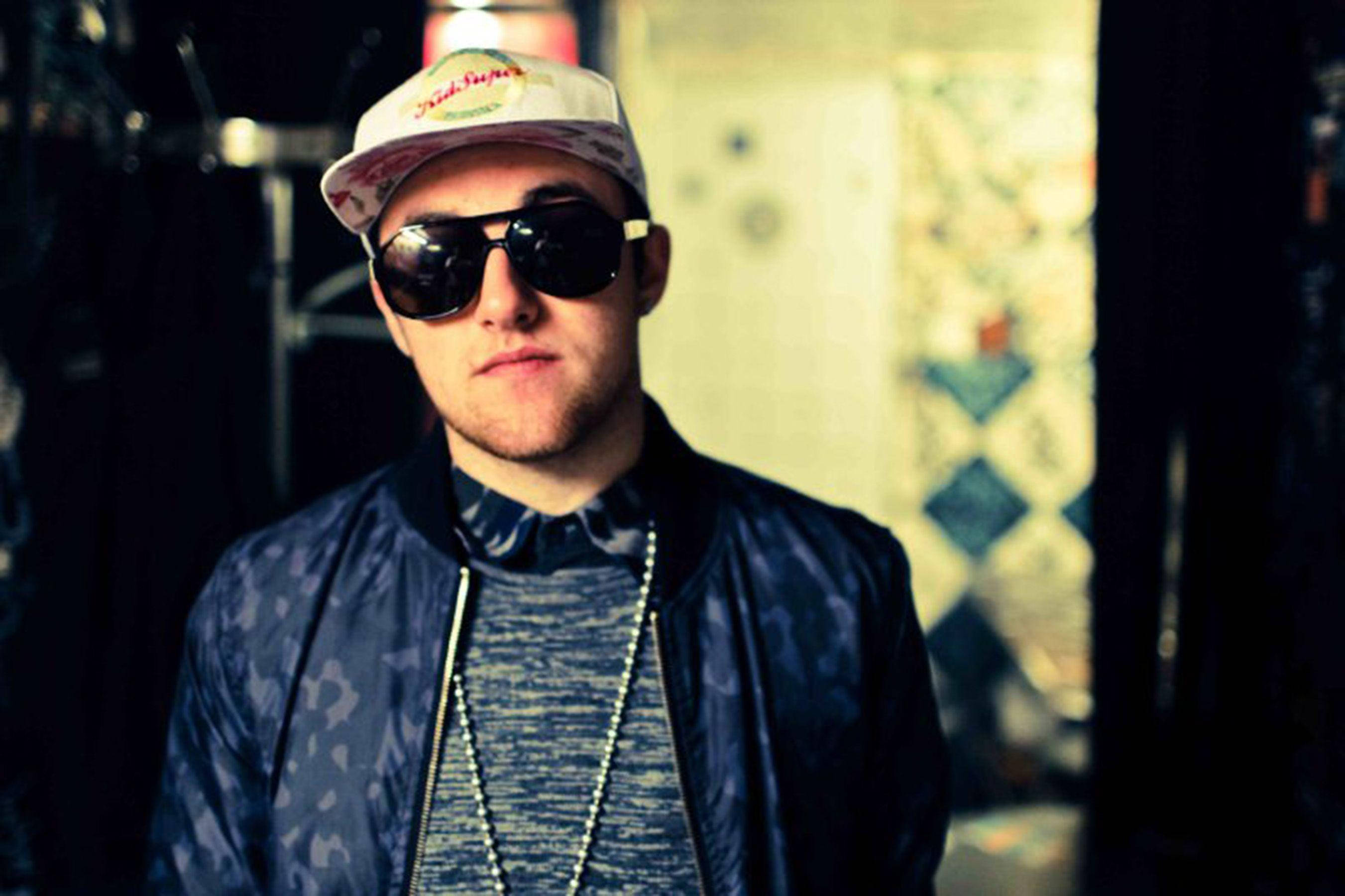 Mac Miller 桌布 壁紙 — 一名戴著圖案帽子和太陽眼鏡的年輕男子在光線昏暗、牆上鋪滿彩色瓷磚的房間裡，凝視著鏡頭。