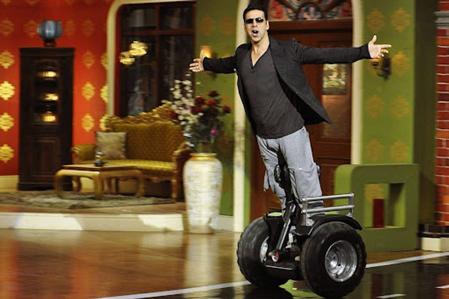 Akshay Kumar 桌布 壁紙 — 一名戴著太陽眼鏡、穿著休閒服裝的男子，在裝飾華麗的電視攝影棚場景中，騎著兩輪電動滑板車保持平衡。