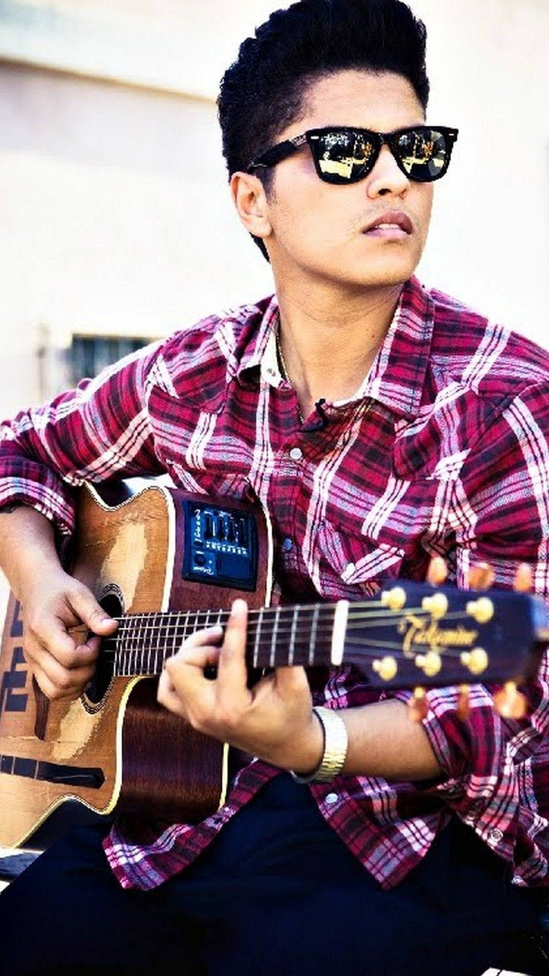 Bruno Mars 桌布 壁紙 — 一名戴著太陽眼鏡、身穿紅色格紋襯衫的年輕男子在戶外明媚陽光下彈奏木吉他。