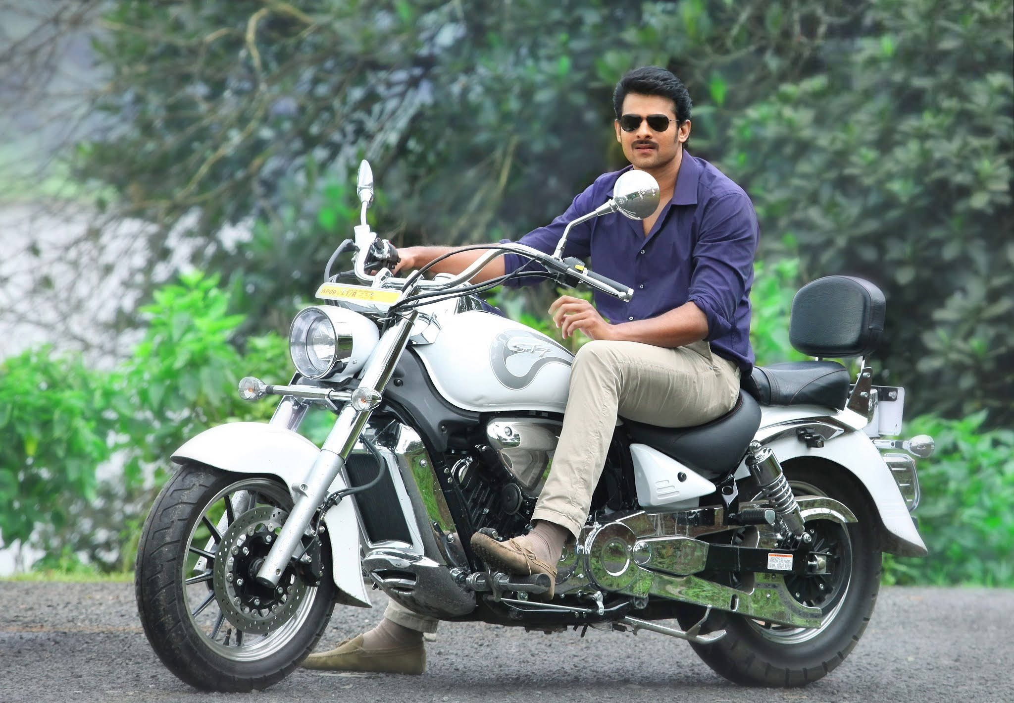 Prabhas Mirchi 桌布 壁紙 — 一名戴著太陽眼鏡、身穿紫色襯衫的男子自信地坐在一輛停在碎石路上的白色巡航摩托車上，周圍是茂密的綠色植物。