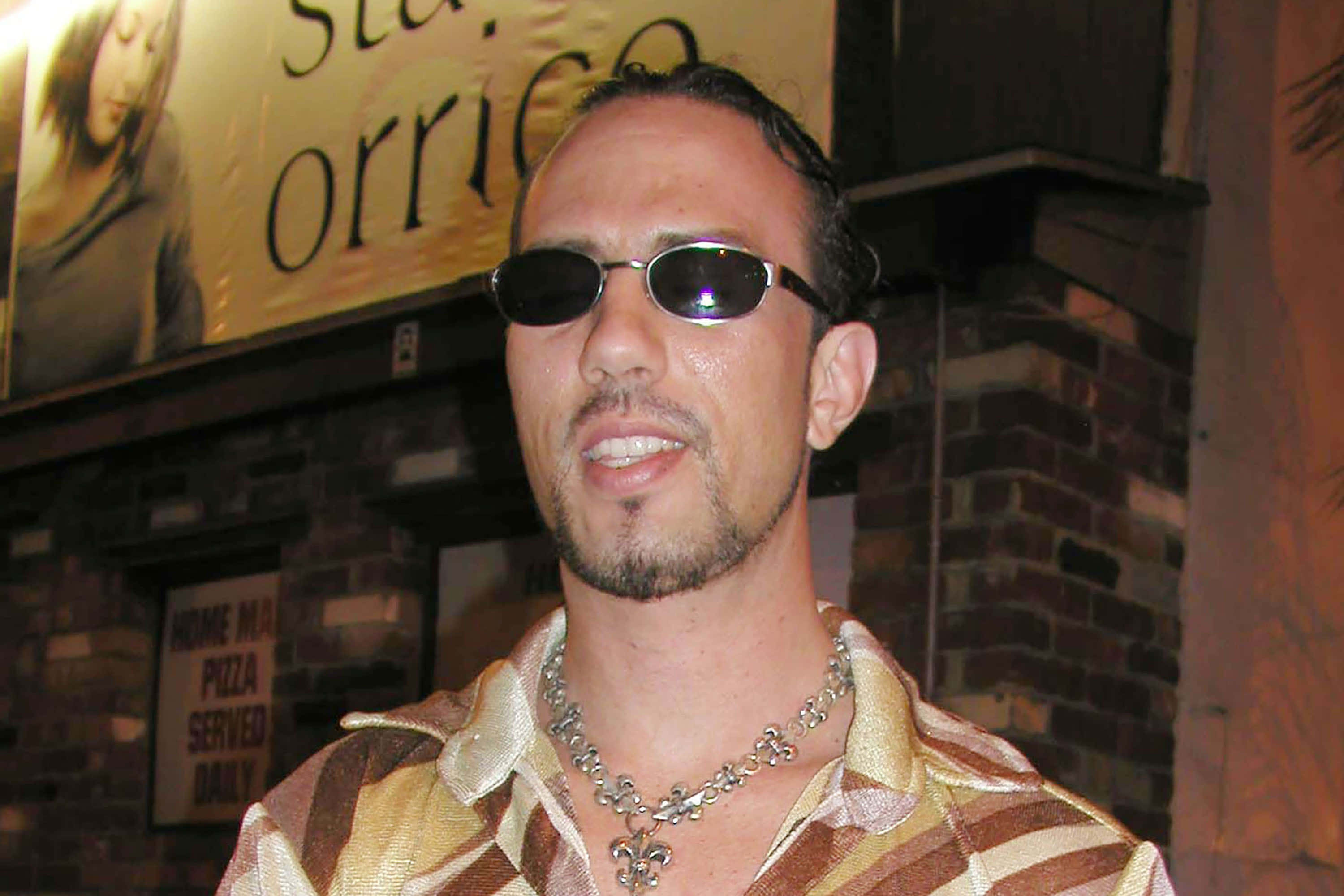 WWE 桌布 壁紙 — 一名戴著太陽眼鏡和圖案襯衫的男子在磚牆前微笑，牆上有披薩招牌。