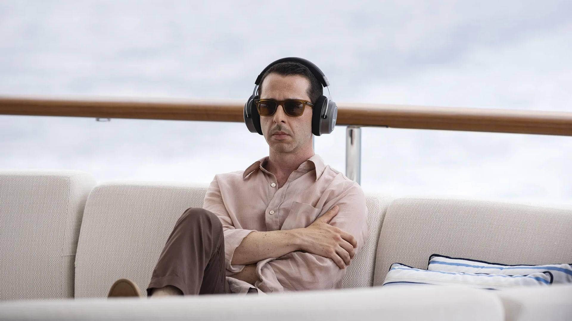 Succession 桌布 壁紙 — 一名戴著太陽眼鏡和耳機的男子，放鬆地坐在遊艇沙發上，俯瞰陰天下的平靜海面。