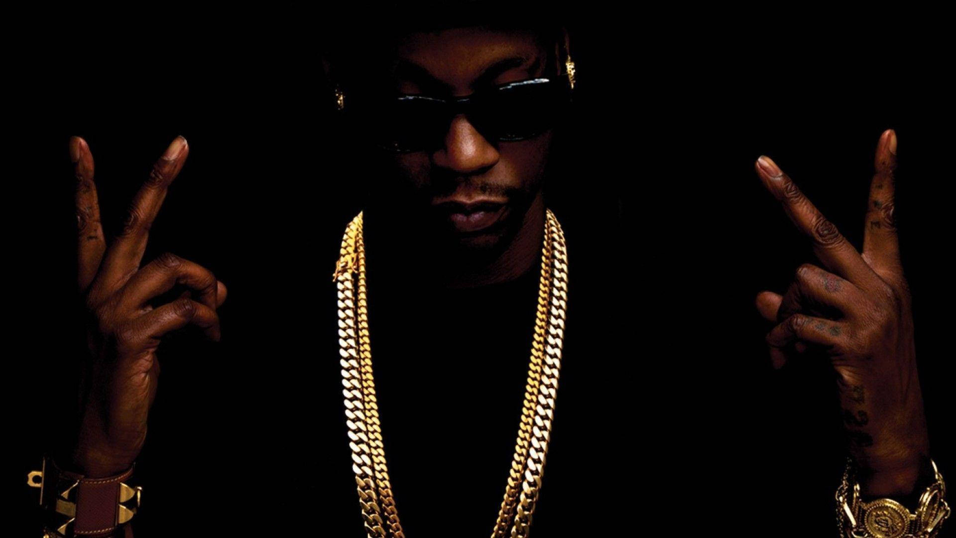2 Chainz 桌布 壁紙 — 一名戴著太陽眼鏡和金鏈的男子在黑色背景上以戲劇性燈光做出和平手勢。