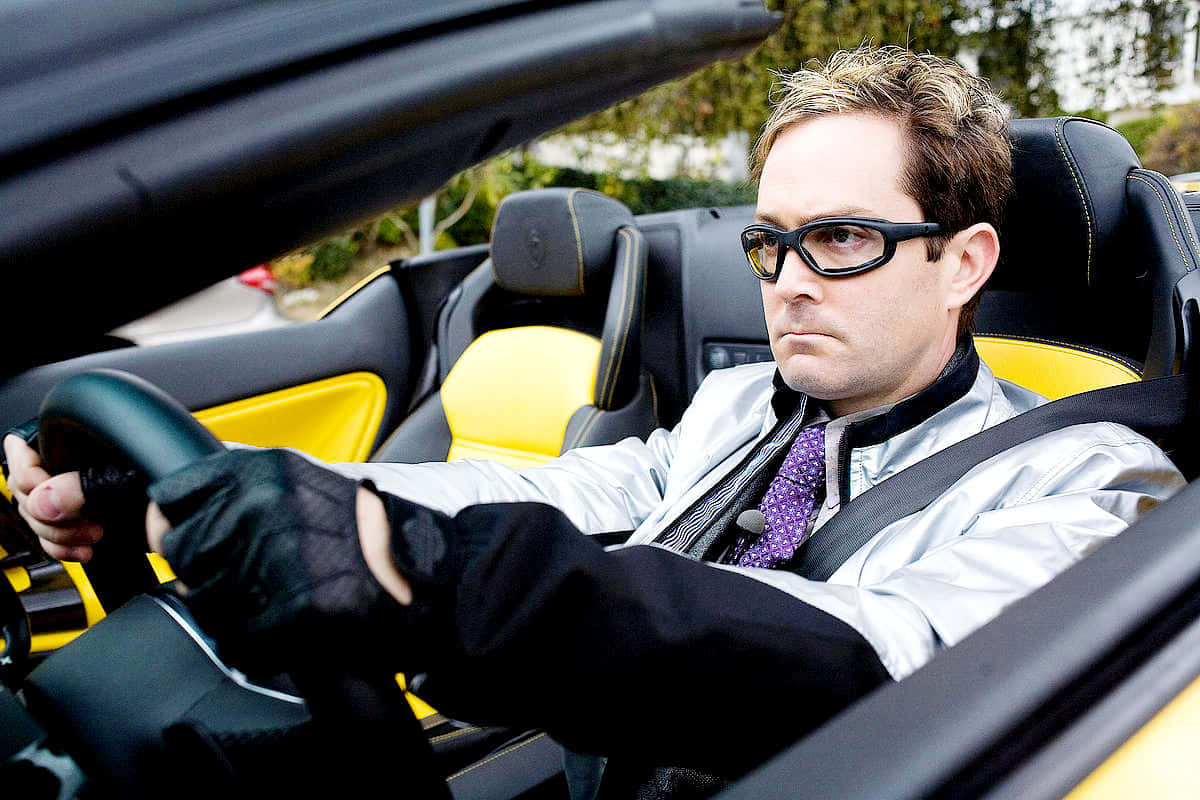 thomas lennon 桌布 壁紙 — 一名戴著太陽眼鏡和銀色外套的男子握著一輛黃黑雙色敞篷跑車的方向盤。