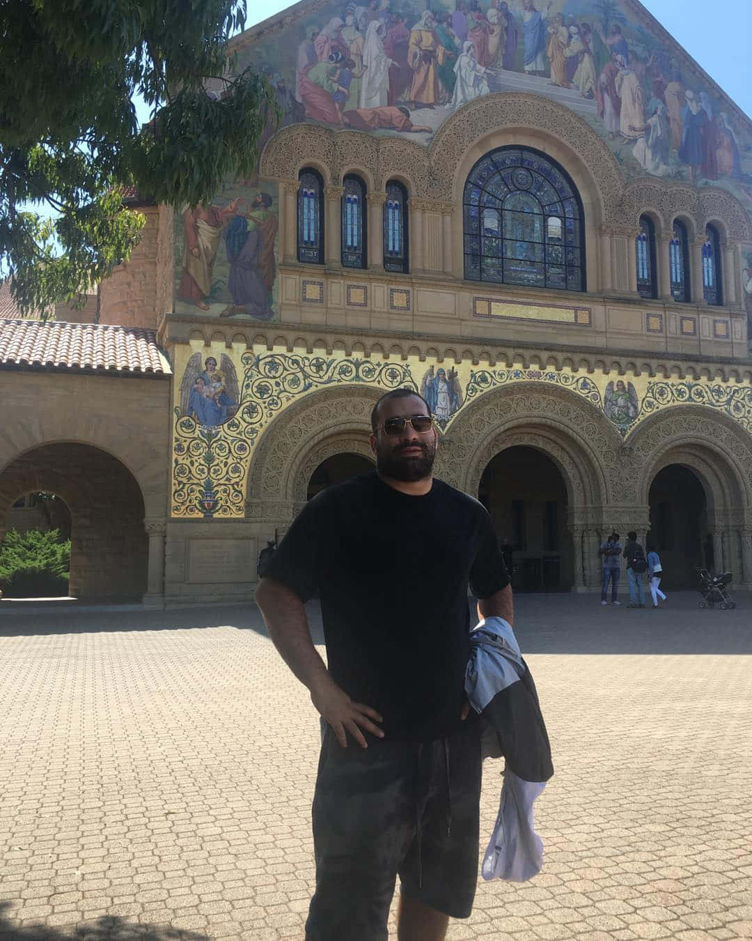 教堂 桌布 壁紙 — 一名戴著太陽眼鏡的男子自信地站在一座宏偉的建築前，建築物裝飾著色彩繽紛的宗教壁畫和拱形窗戶，在明亮的陽光下。