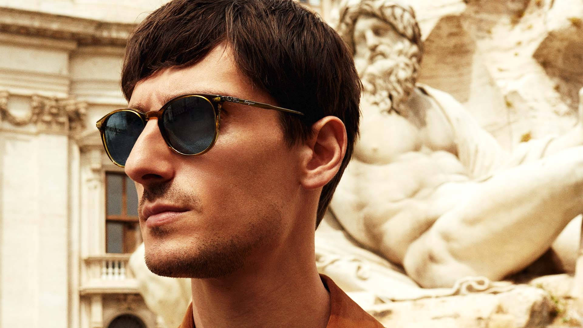 Ferragamo 桌布 壁紙 — 一名戴著時尚太陽眼鏡的男子，在陽光明媚的歐洲廣場上，若有所思地凝視著一座古典大理石雕像。