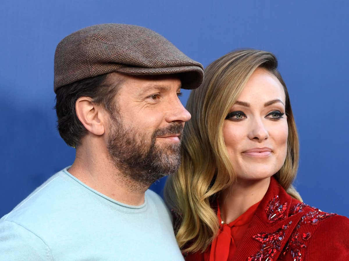 jason sudeikis 桌布 壁紙 — 一名戴著棕色平頂帽的男人和一名穿著紅色裝飾上衣的女人在純藍色背景前緊密擺姿勢。