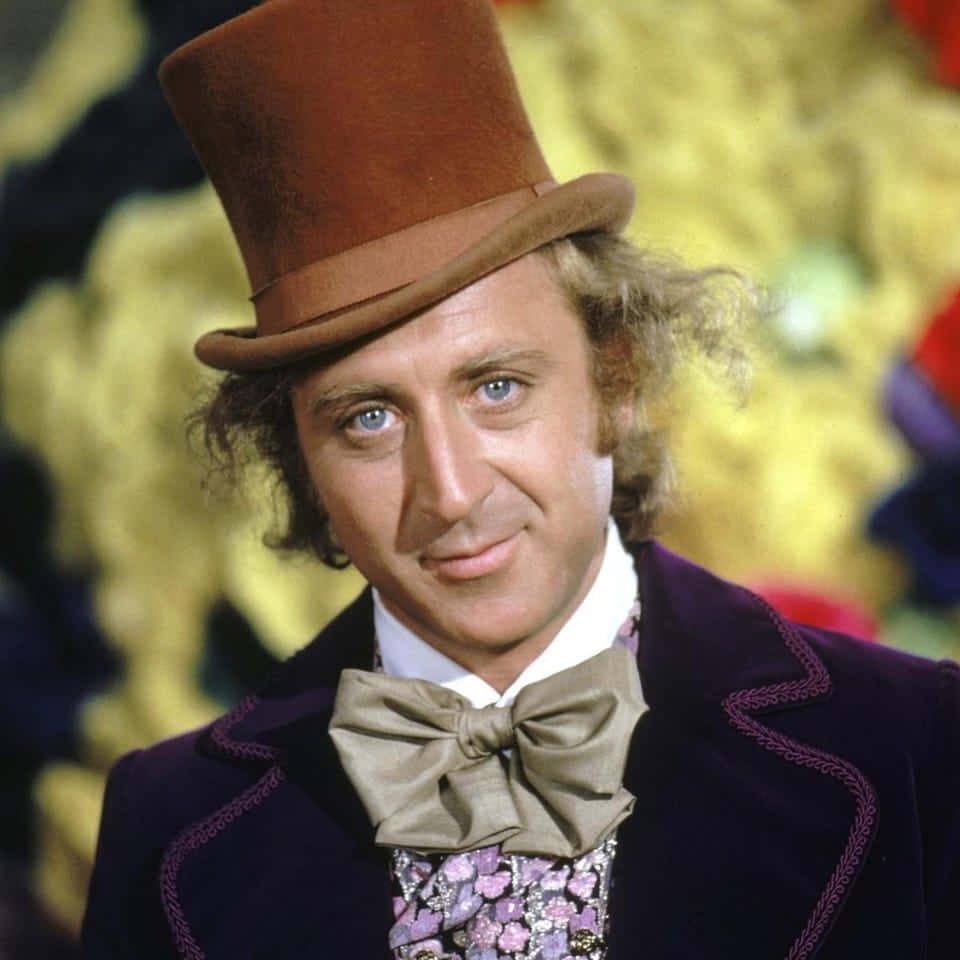 Willy Wonka 桌布 壁紙 — 一名戴著棕色高頂帽和紫色天鵝絨外套的男子，在色彩繽紛的模糊背景前溫暖地微笑。