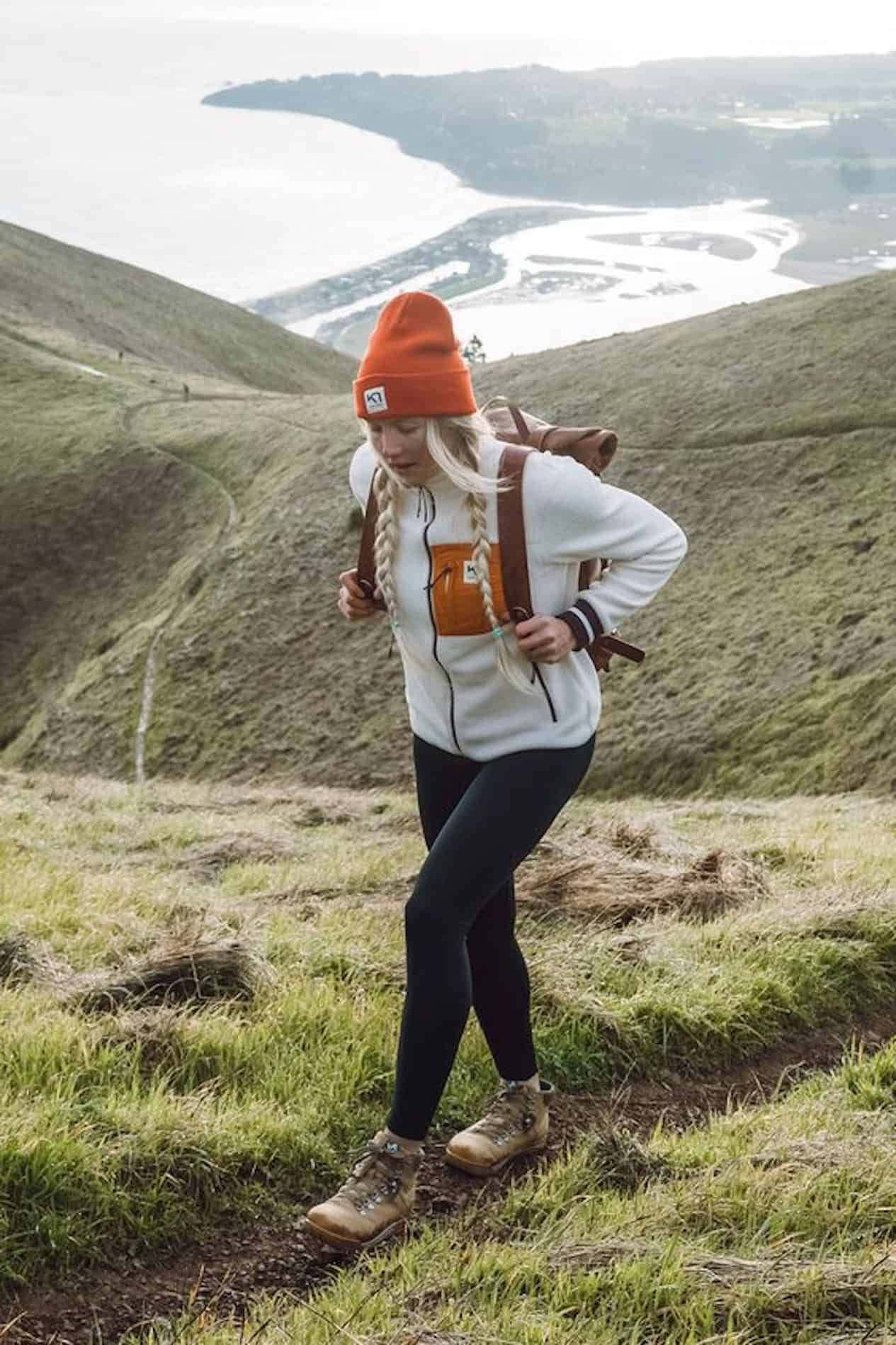 Winter Hiking 桌布 壁紙 — 一名戴著橙色毛帽、身穿白色連帽上衣的女子，在俯瞰蜿蜒海岸灣的草地上健行。