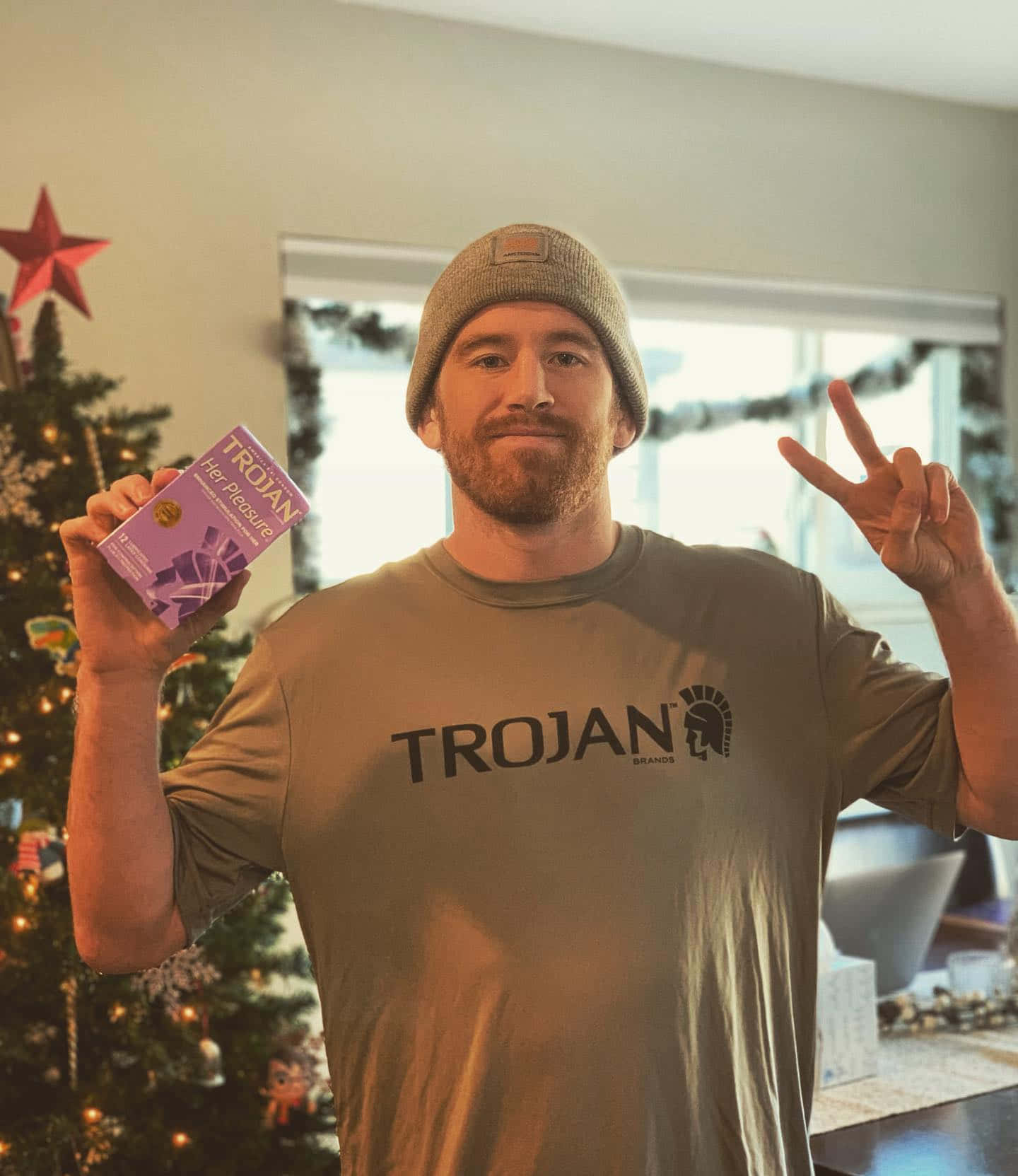 聖誕樹動畫 桌布 壁紙 — 一名戴著毛帽、身穿Trojan T恤的男子微笑著，手持Trojan Her Pleasure盒子，在點亮的聖誕樹旁做出和平手勢。