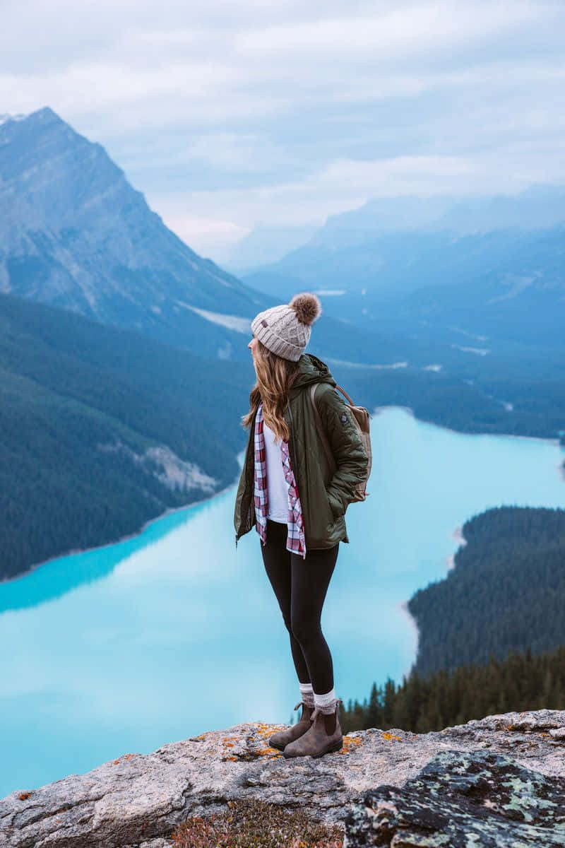 Winter Hiking 桌布 壁紙 — 一名戴著毛帽和外套的女子站在岩石懸崖上，俯瞰著迷霧山巒之間令人驚嘆的碧綠湖泊。