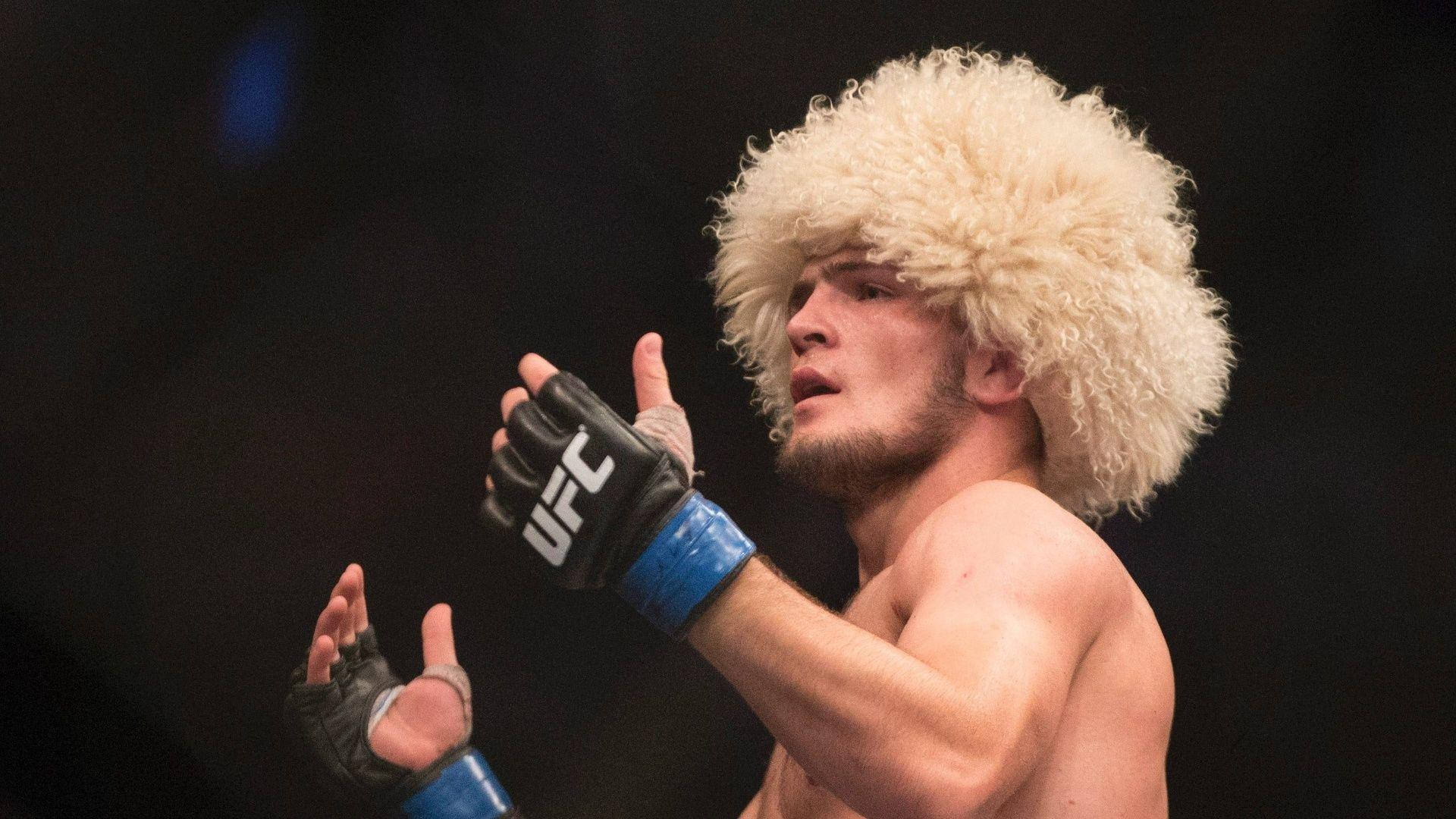 UFC 桌布 壁紙 — 一名戴著毛茸茸白色帽子和UFC手套的拳手在黑暗的競技場中激烈地做手勢。