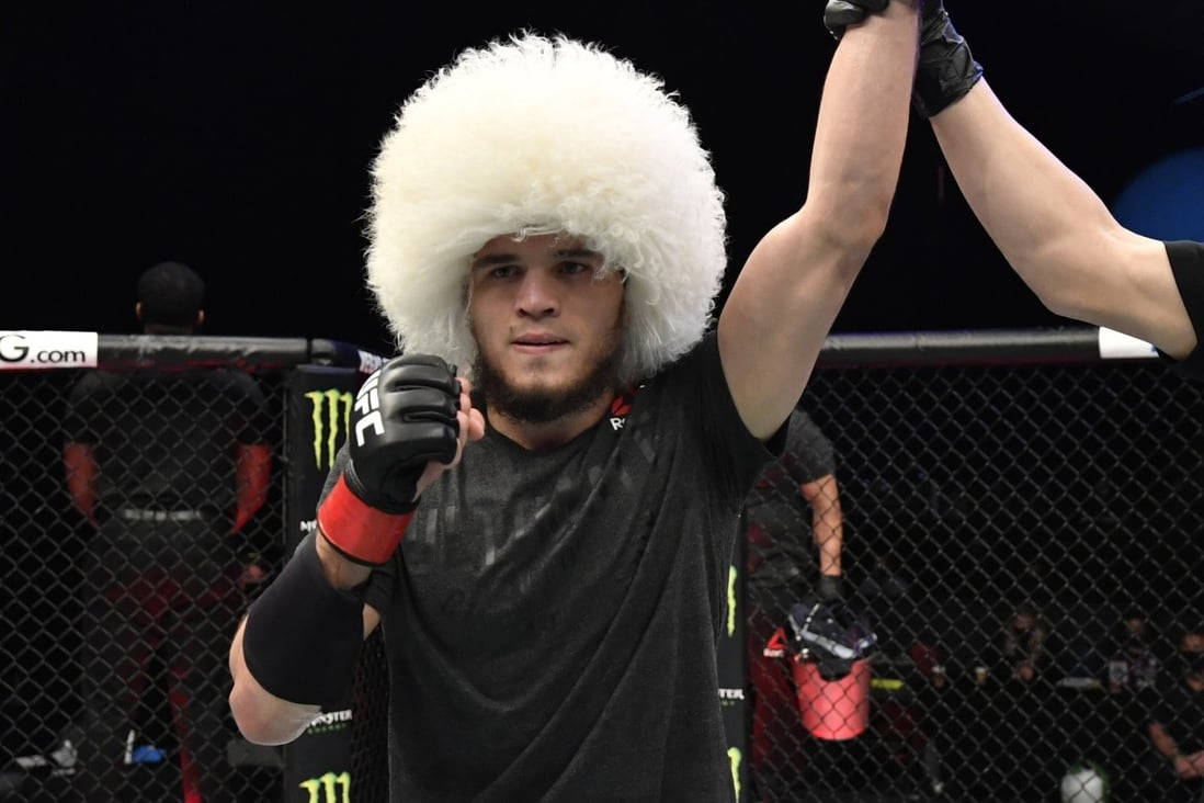 UFC 桌布 壁紙 — 一名戴著毛茸茸白色帽子的戰士在明亮的場館燈光下，於UFC八角籠內舉起手臂慶祝勝利。