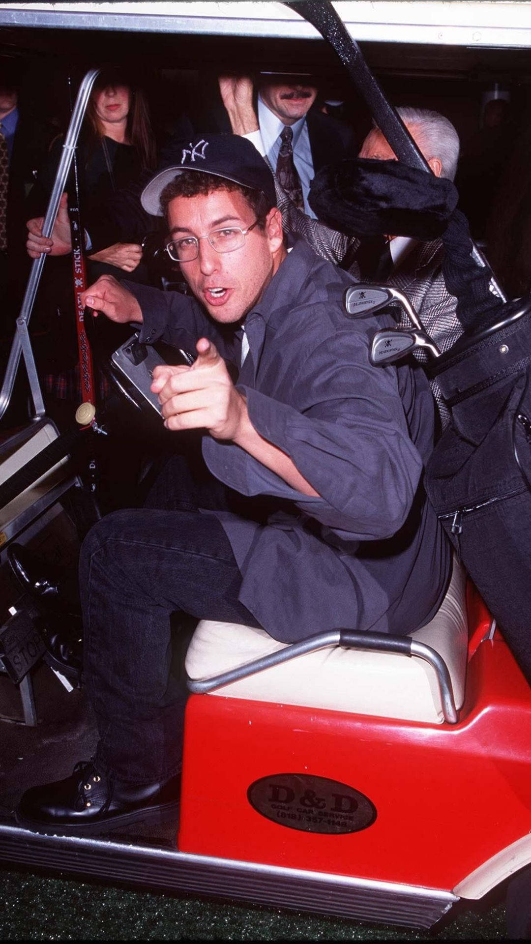 Adam Sandler 桌布 壁紙 — 一名戴著洋基隊帽和眼鏡的男子，在紅色高爾夫球車上頑皮地指著，周圍有許多人。