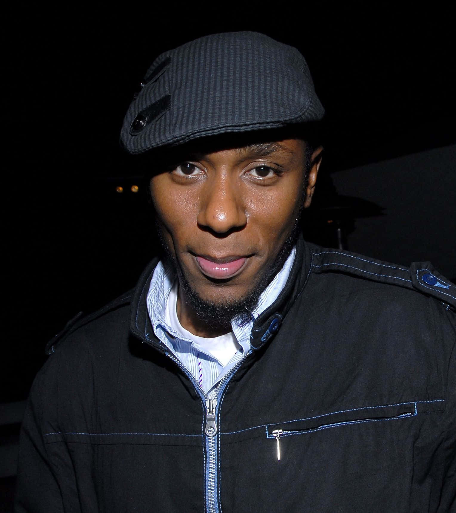 Yasiin Bey 桌布 壁紙 — 一名戴著深色細條紋帽子和黑色外套的男子，在昏暗背景中帶著遠方燈光，溫柔地微笑。