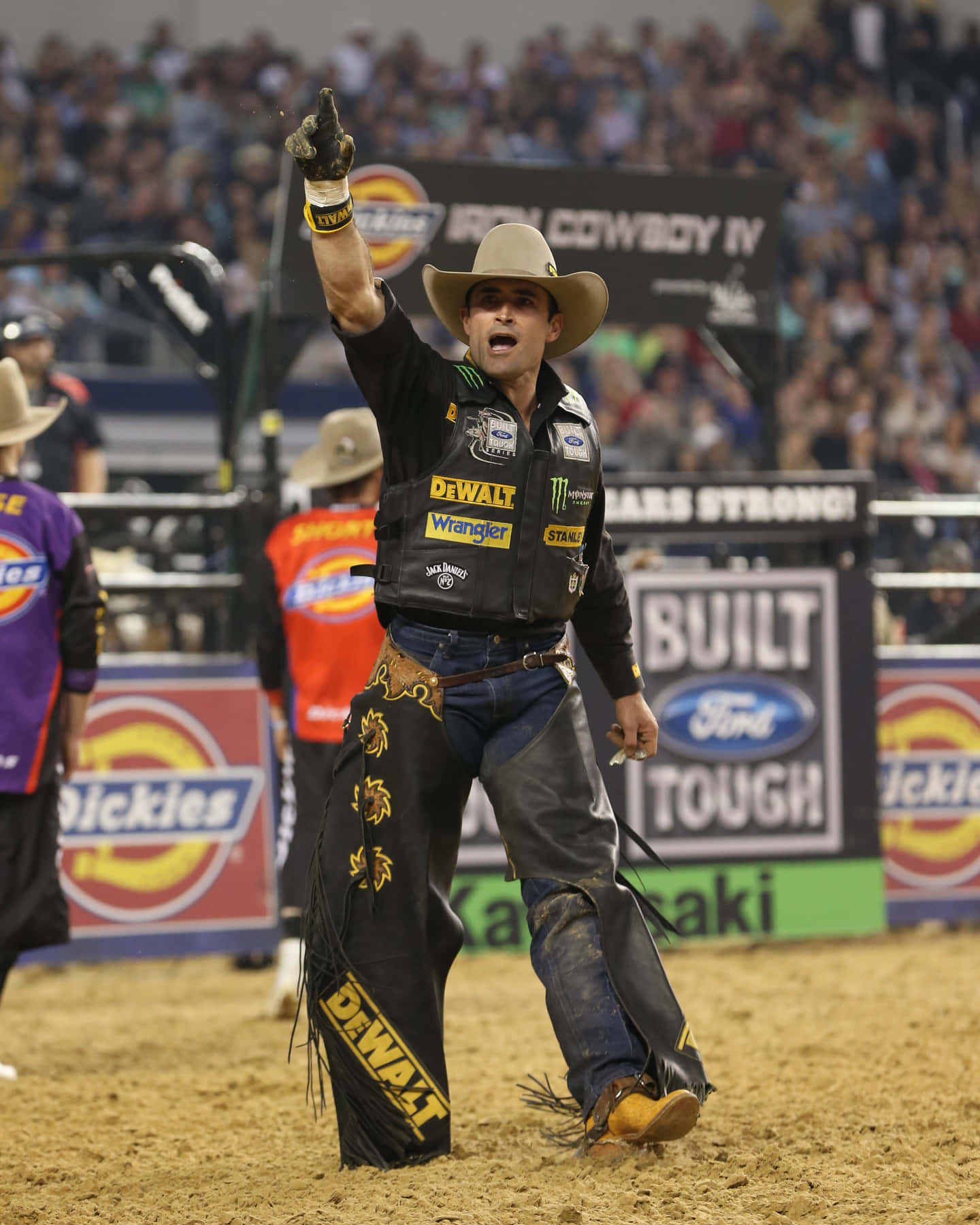 Bull Rider 桌布 壁紙 — 一名戴著牛仔帽、身穿品牌背心的鬥牛騎士在牛仔競技場高舉單臂慶祝勝利。