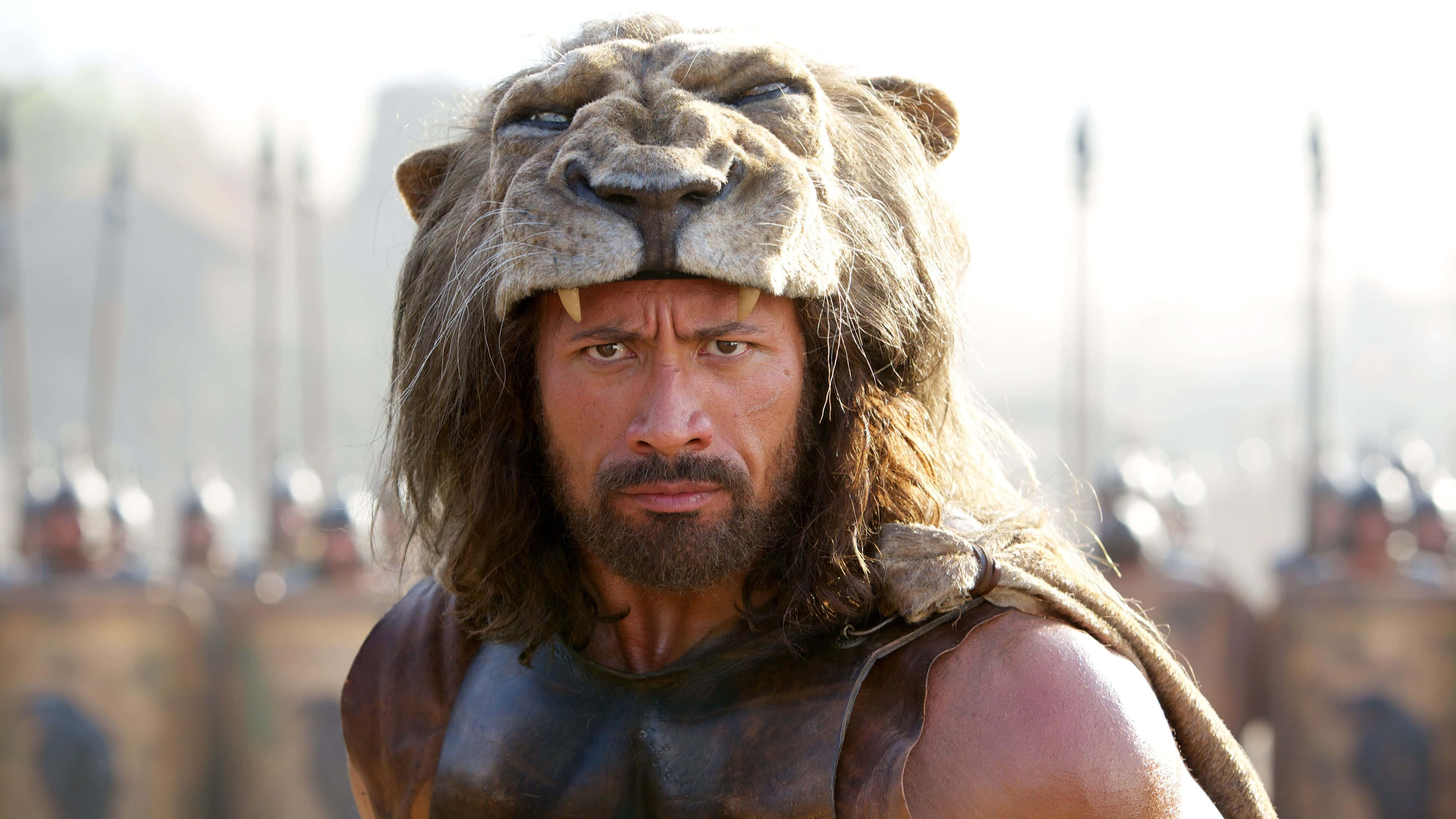 海克力斯 桌布 壁紙 — 一名戴著獅頭頭盔的戰士，身後有模糊的士兵和長矛，目光炯炯地向前凝視。