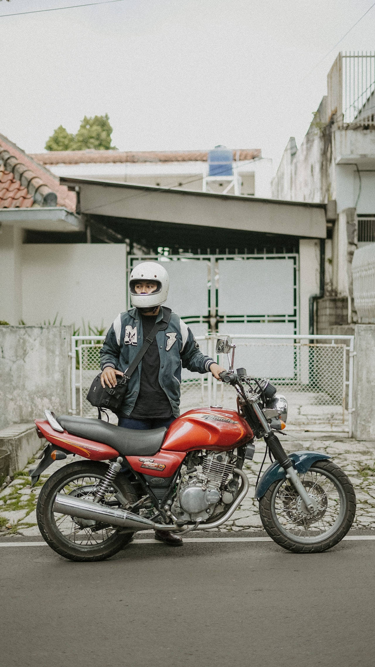 bike riding 桌布 壁紙 — 一名戴著白色安全帽的人站在一輛停放在寧靜住宅街道上的紅藍色機車旁。