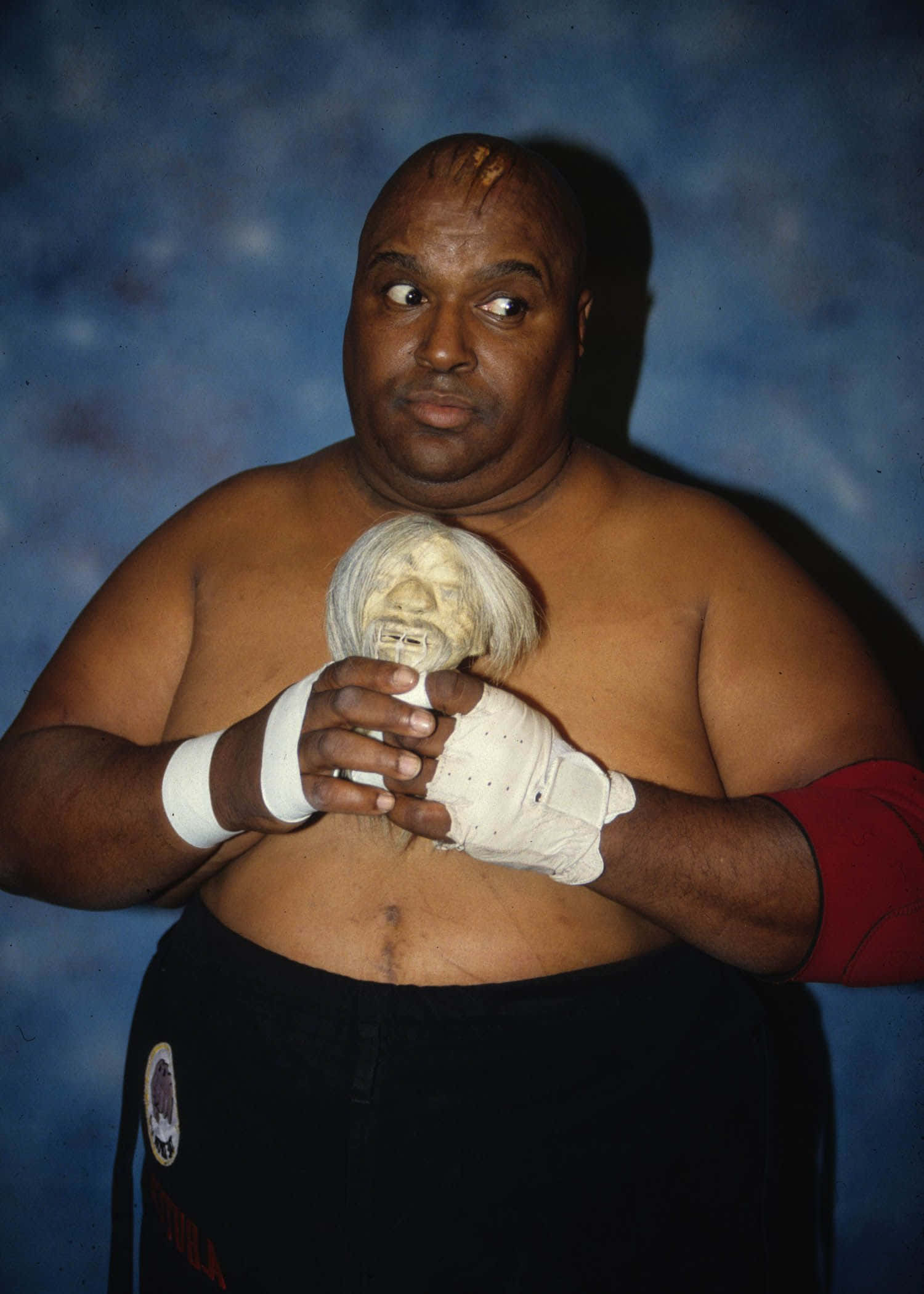 Abdullah The Butcher 桌布 壁紙 — 一名戴著白色手套的摔角手，在帶有紋理的藍色背景前，手持一個怪誕的白色面具。