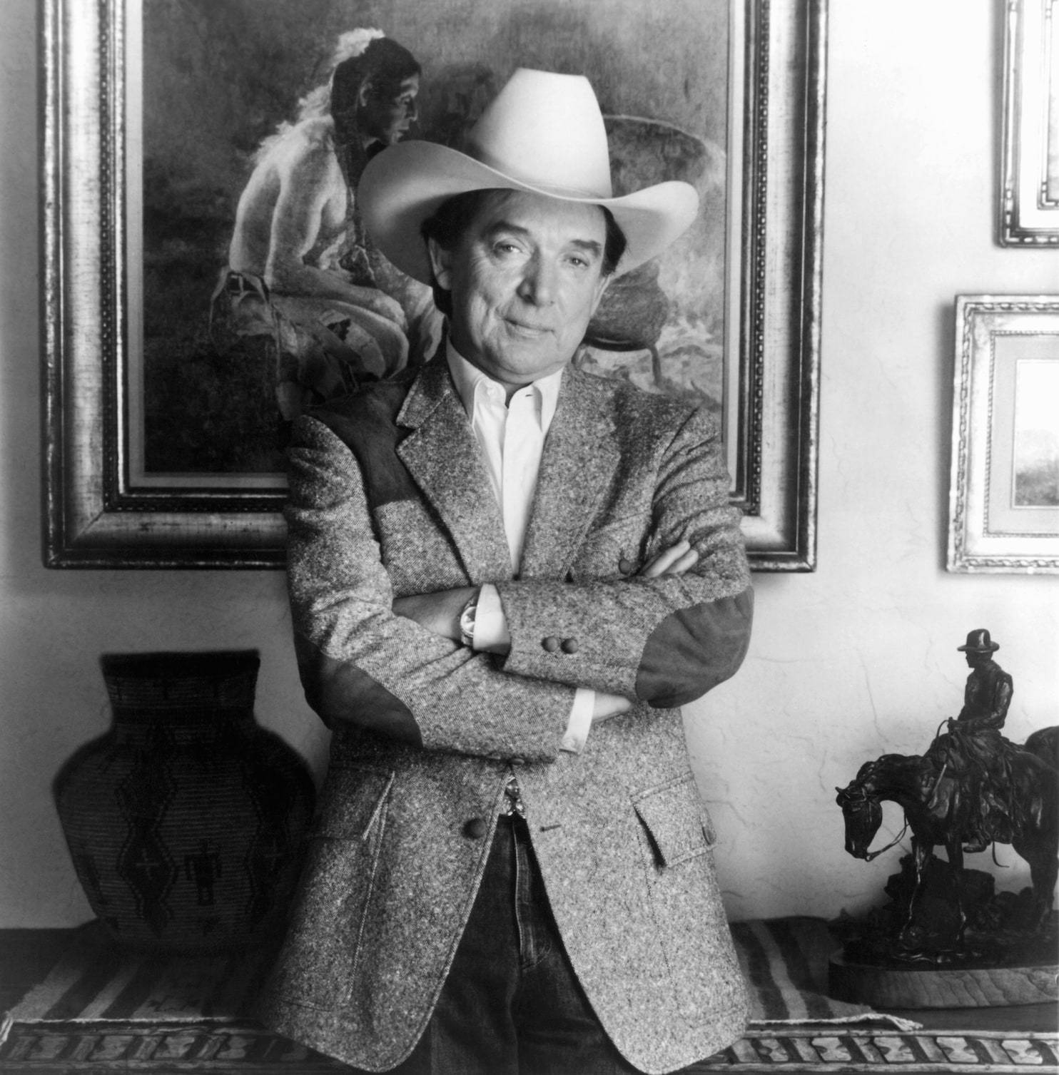 Cowboy 桌布 壁紙 — 一名戴著白色牛仔帽、身穿質感外套的男子，自信地交叉雙臂站在一個裝飾著裱框藝術品和美洲原住民文物的房間裡。