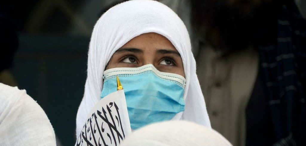 Taliban 桌布 壁紙 — 一名戴著白色頭巾和藍色外科口罩的女子，手持帶有阿拉伯文字的標語牌，抬頭仰望。