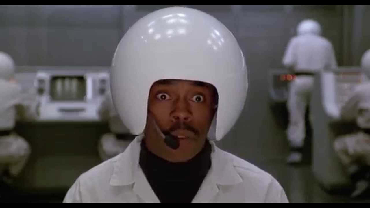 spaceballs 桌布 壁紙 — 一名戴著白色頭盔和實驗室外套的男子在無菌的未來實驗室中睜大眼睛凝視，背景有其他穿著西裝的人物。