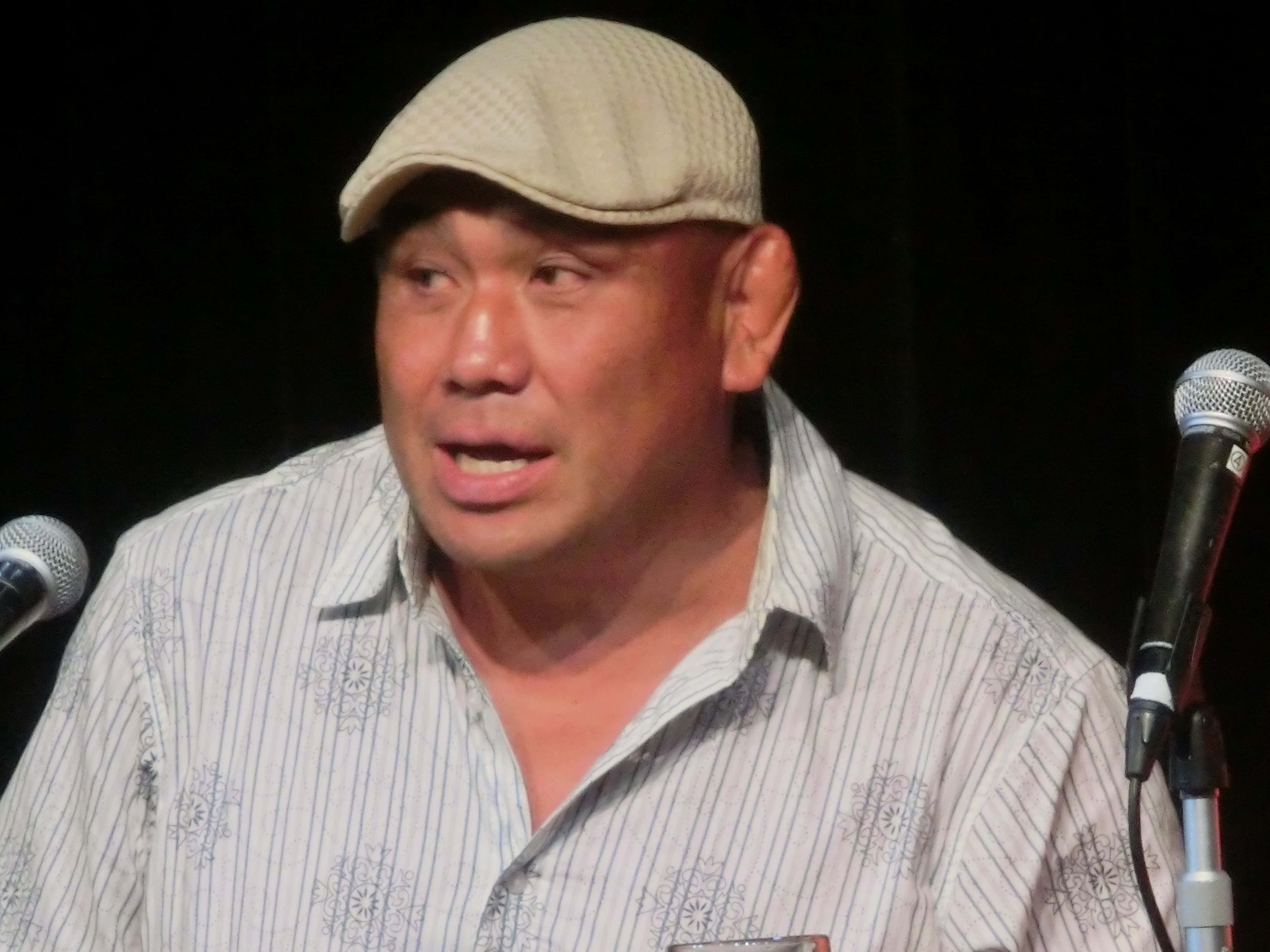 Athlete 桌布 壁紙 — 一名戴著米色帽子和條紋襯衫的男子在深色背景下對著麥克風說話。
