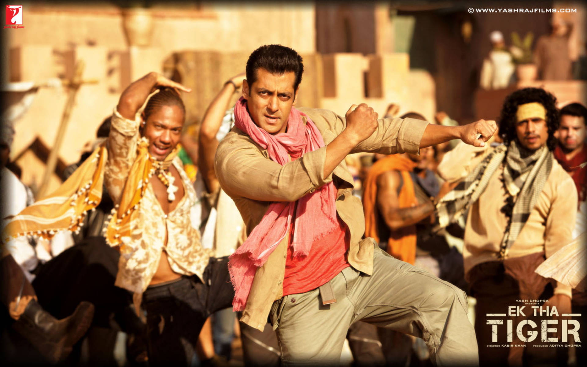 Salman Khan 桌布 壁紙 — 一名戴著粉紅色圍巾的男子在陽光明媚的沙漠場景中擺出強勁的武術姿勢，周圍有歡呼的舞者。