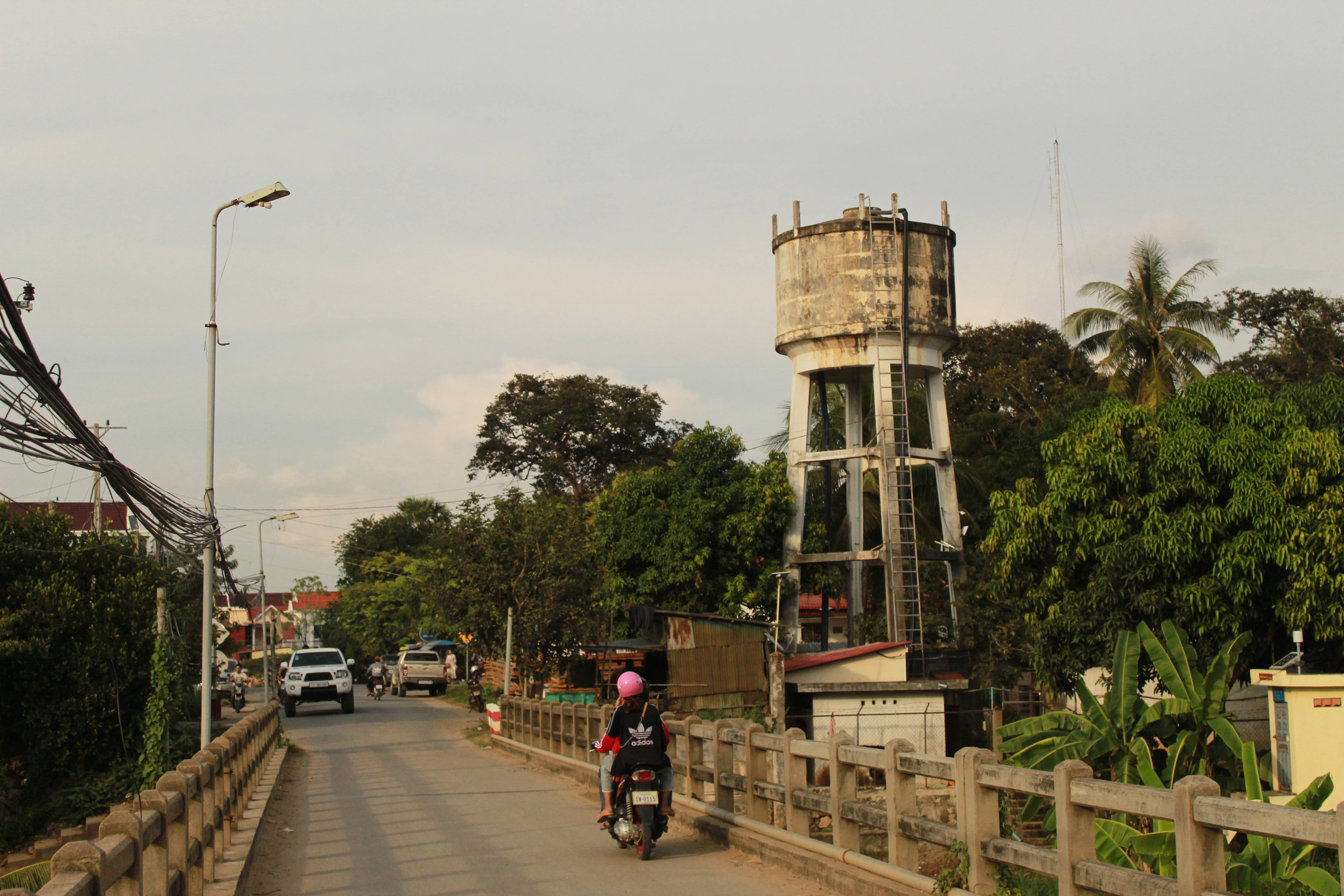 Cambodia 桌布 壁紙 — 一名戴著粉紅色安全帽的機車騎士在陰天背景下，騎過一座橋，背景有風化水塔和茂密的綠樹。