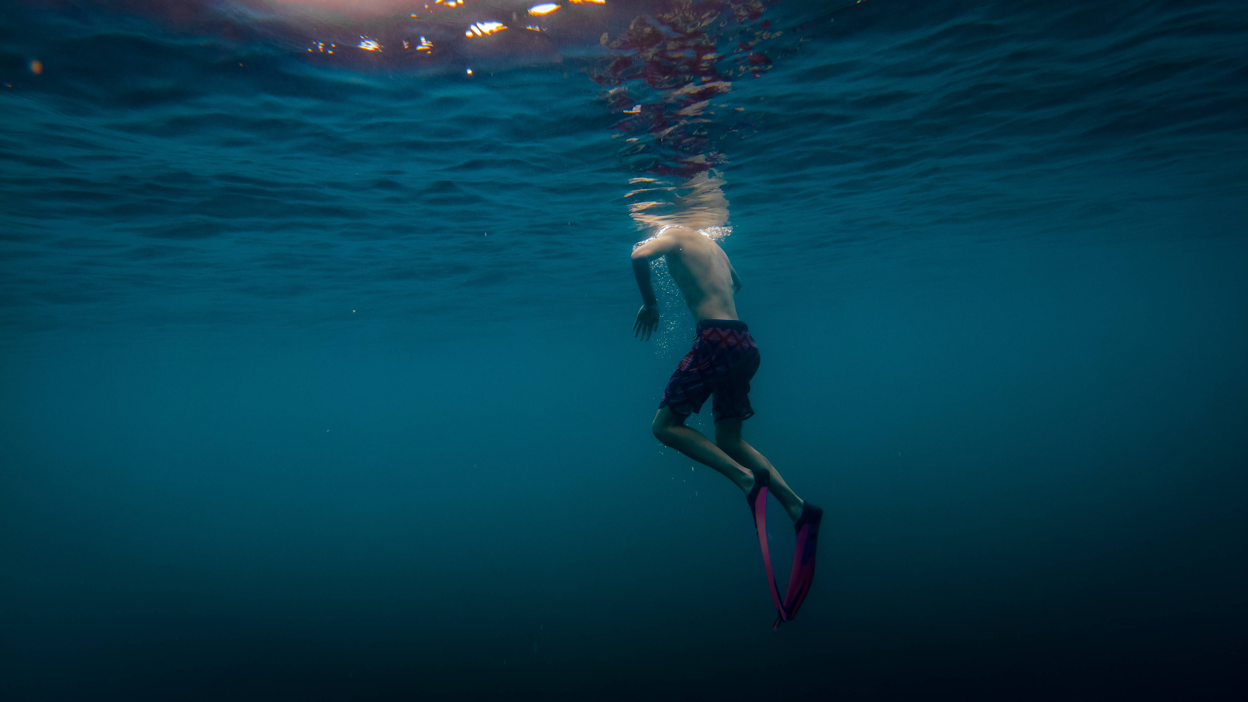 Snorkeling 桌布 壁紙 — 一名戴著粉紅色腳蹼的游泳者潛入水中，朝向深藍色海洋中被陽光照射的水面。