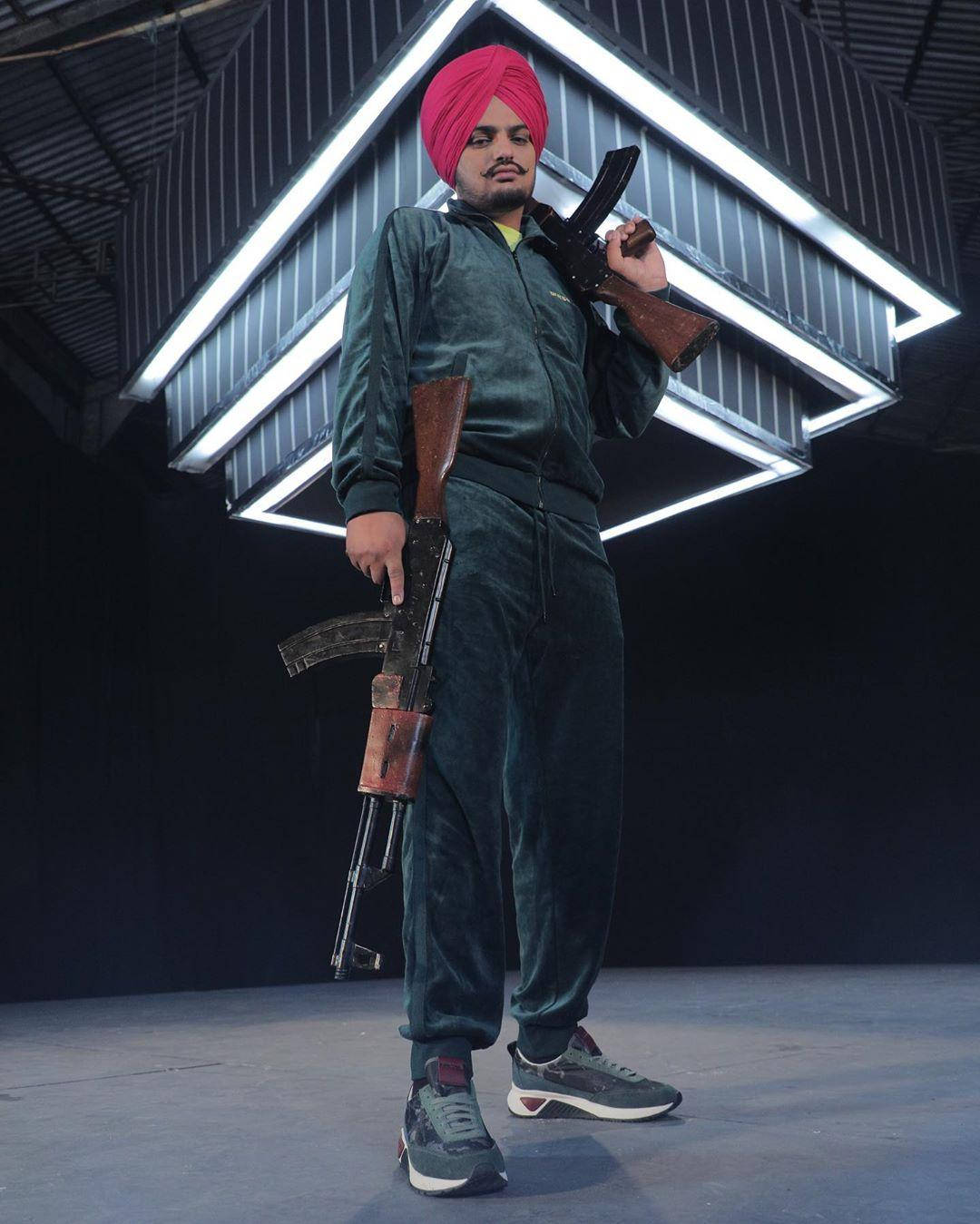 戴粉紅色頭巾、穿綠色天鵝絨西裝的男子在明亮攝影棚燈光下手持步槍。 — 說唱歌手 壁紙