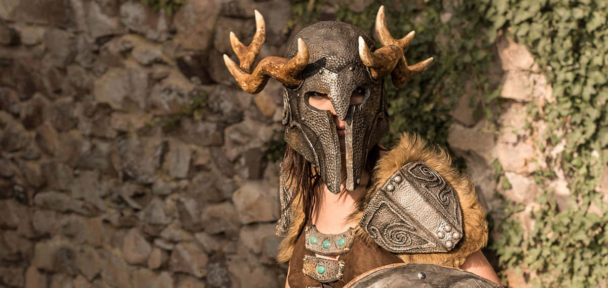 Aela The Huntress 桌布 壁紙 — 一名戴著精緻鹿角頭盔的戰士，站在沐浴在溫暖陽光下的長滿青苔的石牆前。