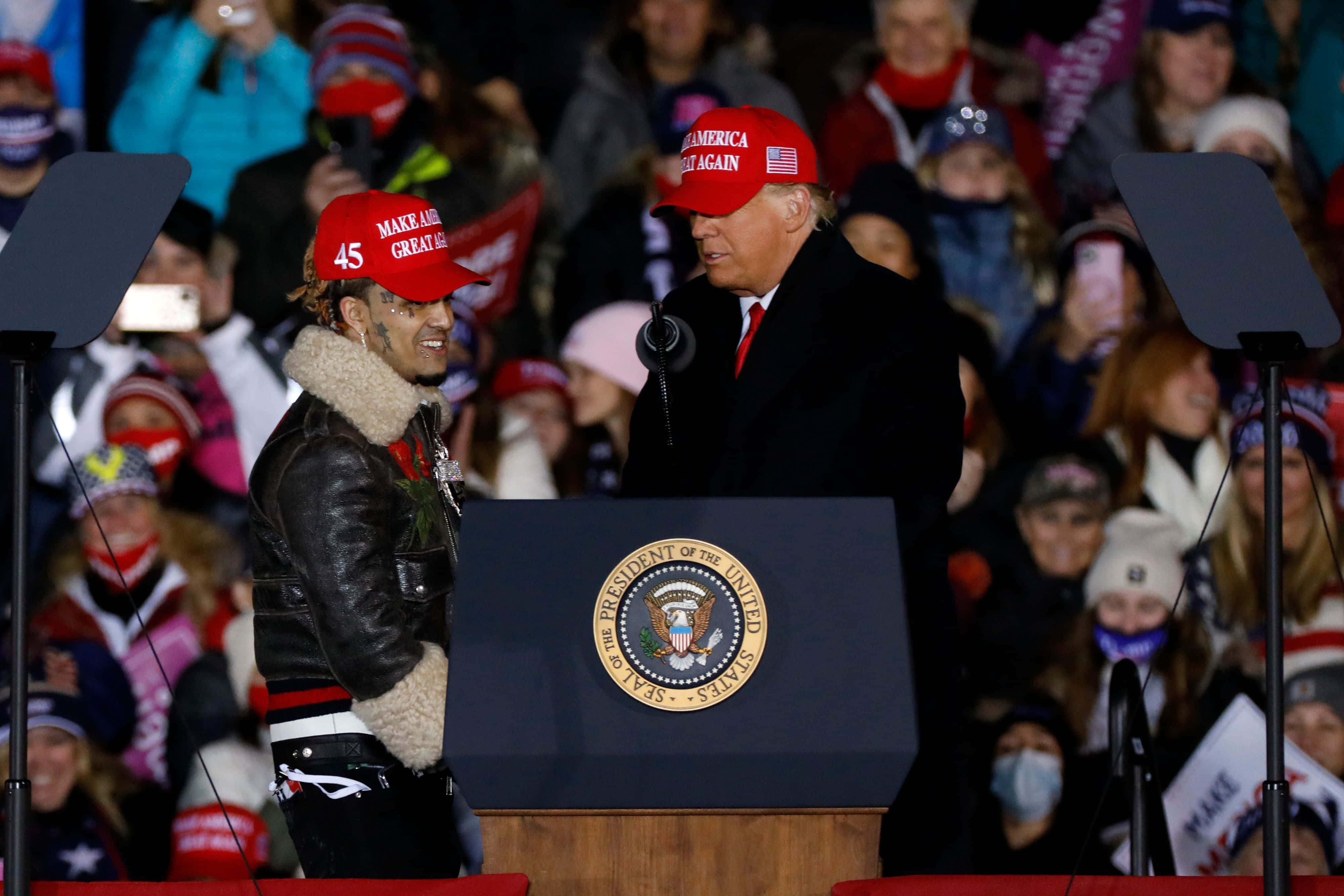 桌布 壁紙 — 一名戴著紅色「Make America Great Again」帽子的男子，在競選集會上站在前總統唐納·川普旁邊，講台上有總統印章。