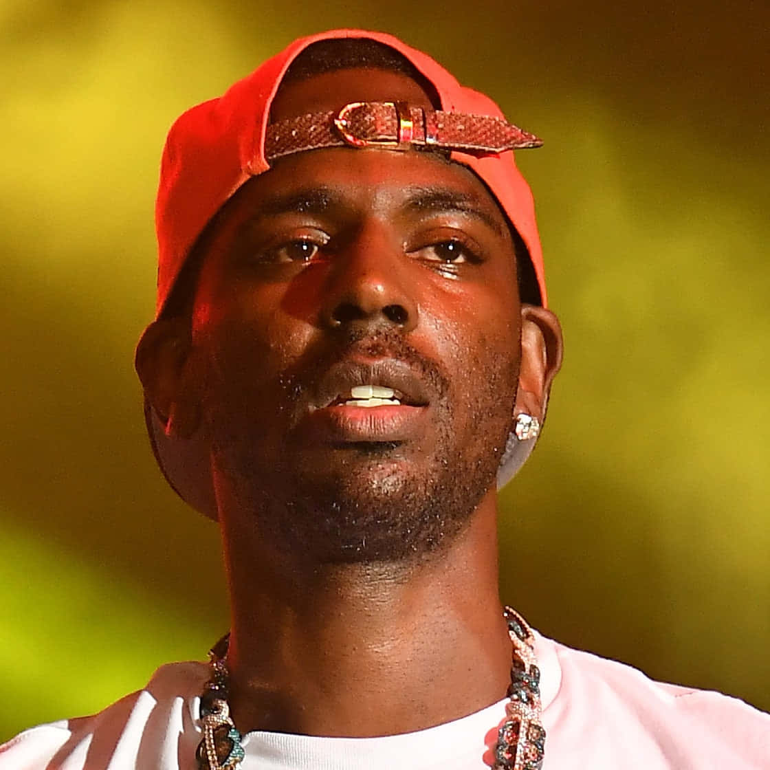 Young Dolph 桌布 壁紙 — 一名戴著紅色帽子和鑽石耳環的男子在發光的黃色背景前的特寫。