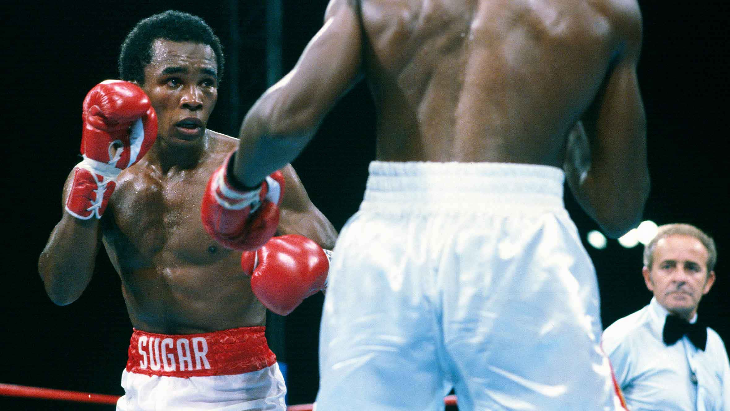 Boxing 桌布 壁紙 — 一名戴著紅色手套、穿著印有「SUGAR」字樣短褲的拳擊手在燈光明亮的擂台上彎腰躲避對手的拳擊。
