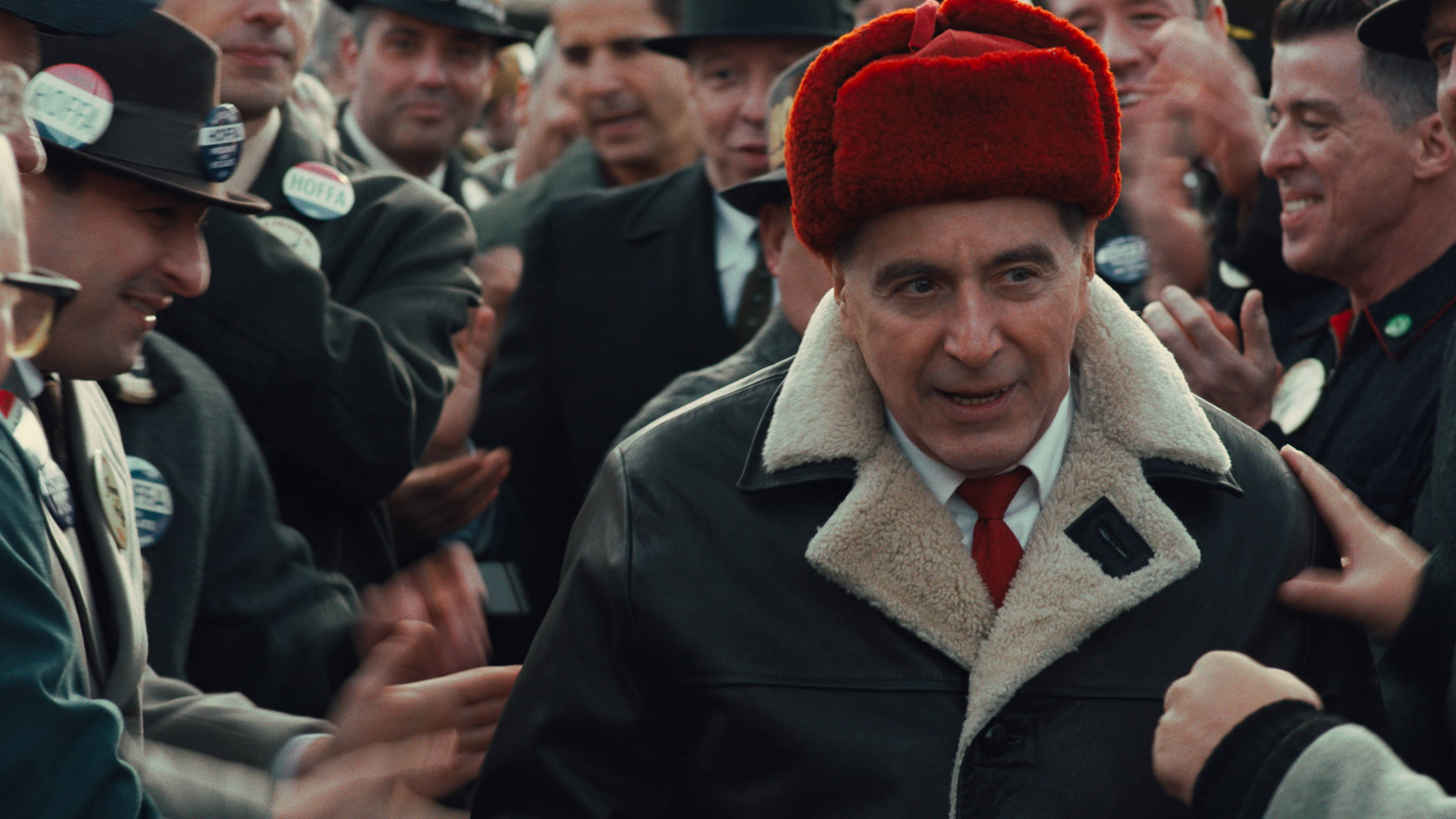 Al Pacino 桌布 壁紙 — 一名戴著紅色毛帽、身穿羊皮大衣的男子，在歡呼的支持者人群中走過，露出溫暖的笑容。