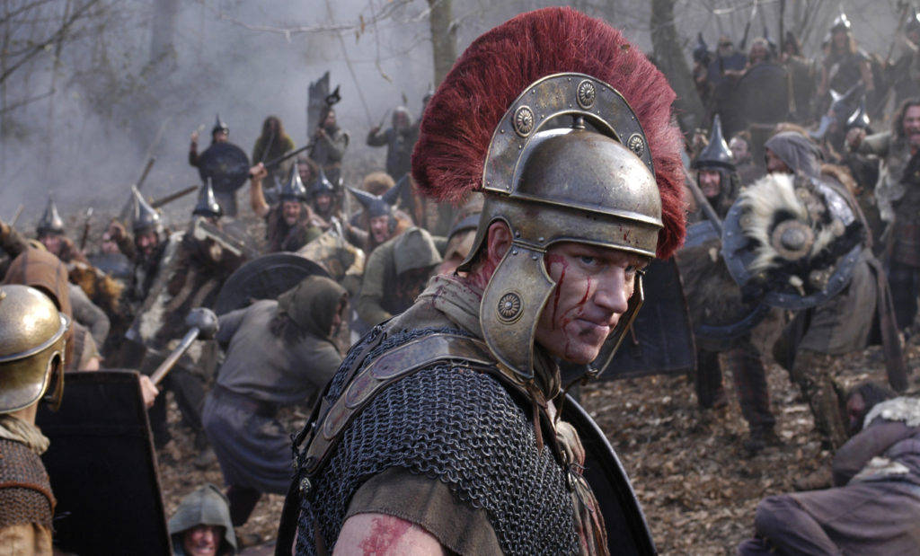rome 2 桌布 壁紙 — 一名戴著紅色羽飾頭盔、滿臉鮮血的羅馬士兵，站在迷霧森林中，被戰士包圍的混亂戰場上。